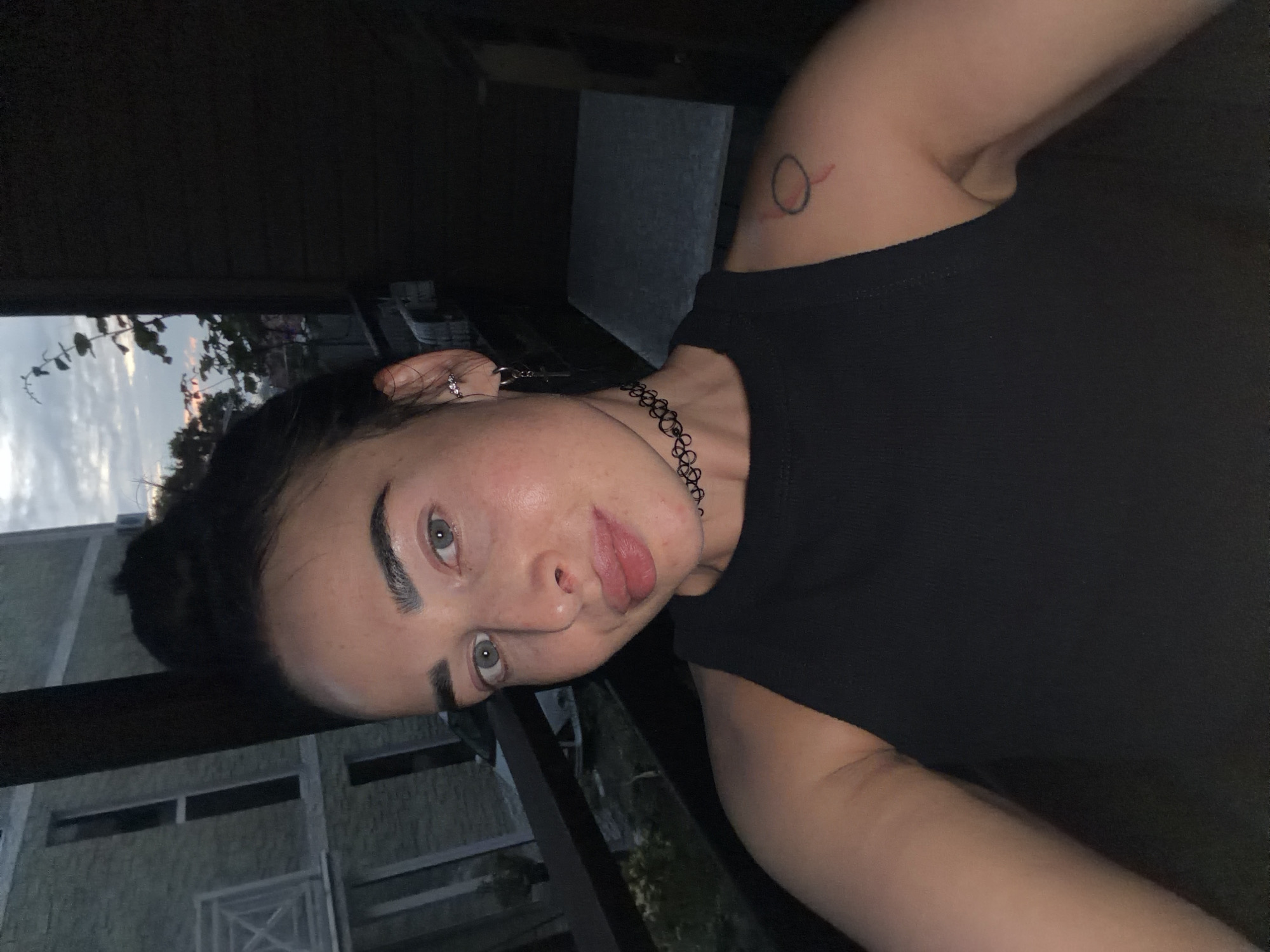 A woman with dark hair pulled into a bun and light blue eyes takes a selfie outdoors. She is wearing a black sleeveless top and a black choker necklace. She has several piercings in her left ear and a small tattoo of a red circle with a line on her right shoulder. In the background, a building with white window frames is visible, as are trees and a cloudy sky at dusk. The image appears to have been taken on a balcony or porch.