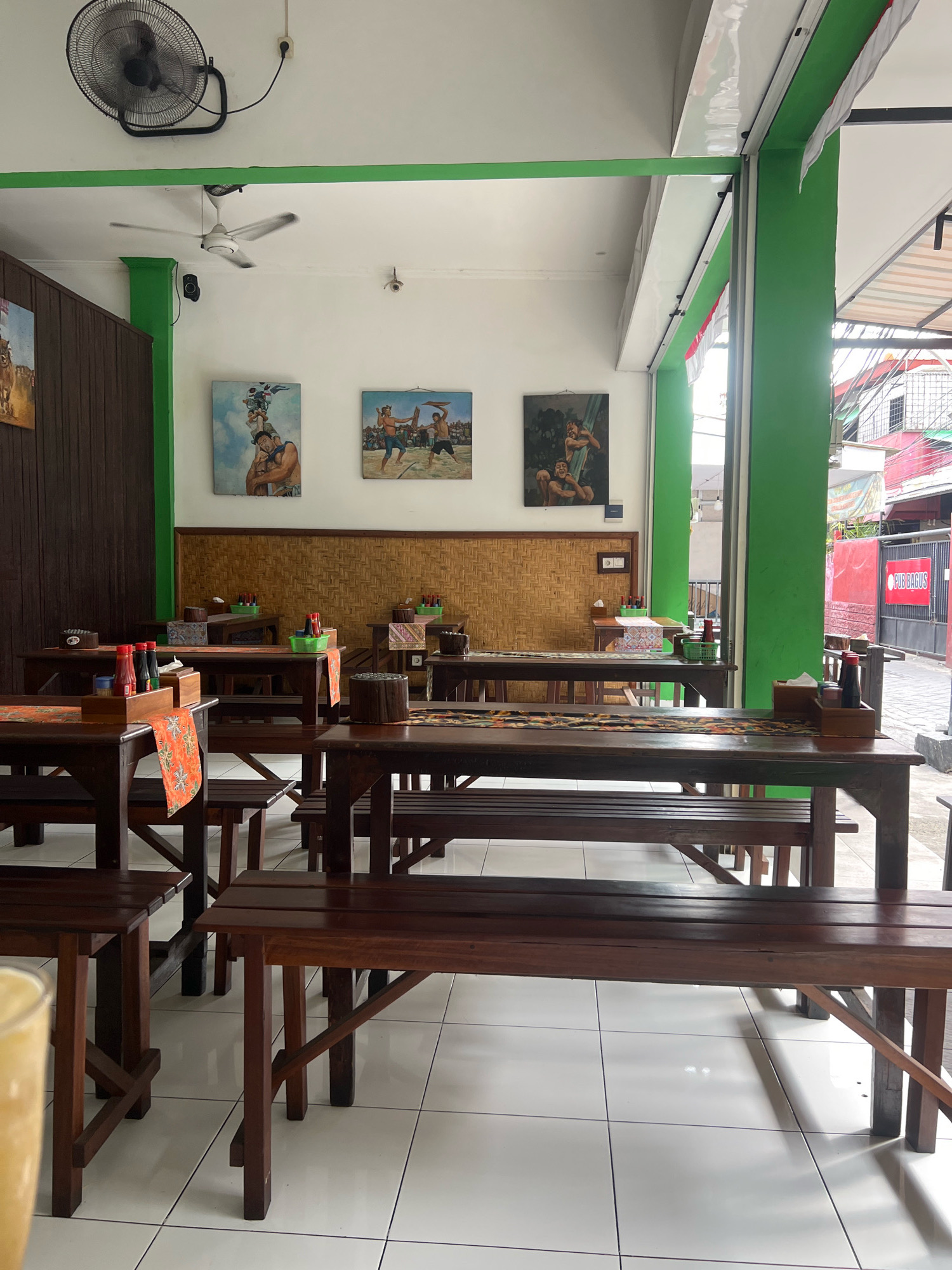 The image depicts the interior of a restaurant in Kuta, Indonesia. The restaurant features dark wooden tables and benches, with a woven bamboo-textured wall in the background. Three paintings of figures in traditional attire hang on the wall. The tables are set with placemats, condiment bottles, and baskets. A ceiling fan is visible on the ceiling, along with a second, smaller fan. The restaurant has bright green trim around the ceiling and window frames. The open doorway reveals a street scene with buildings and signage, including a red building with "PUB BAGUS" visible. The floor is tiled with white square tiles.