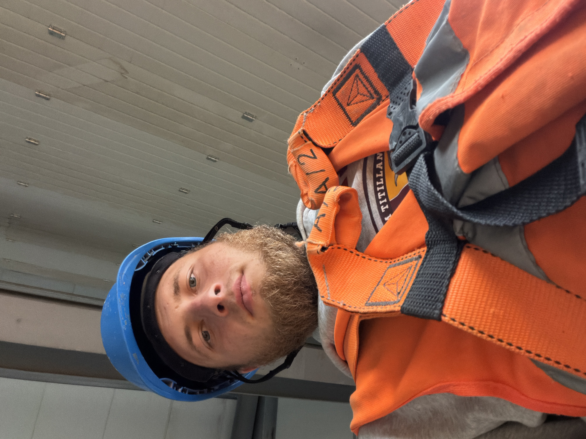 A low-angle, medium shot captures a young, white man wearing a blue hard hat and an orange safety vest. His facial hair is full and light brown, and he looks directly into the camera with a neutral expression. The background consists of a light-colored, corrugated ceiling. The safety vest features black straps and reflective detailing, with a logo that appears to be a triangle within a square, and text reading "A/2" and "TITILLAN". The image is likely taken indoors, possibly on a construction site or industrial facility, though no specific identifying markers of Janowice, Poland are visible.