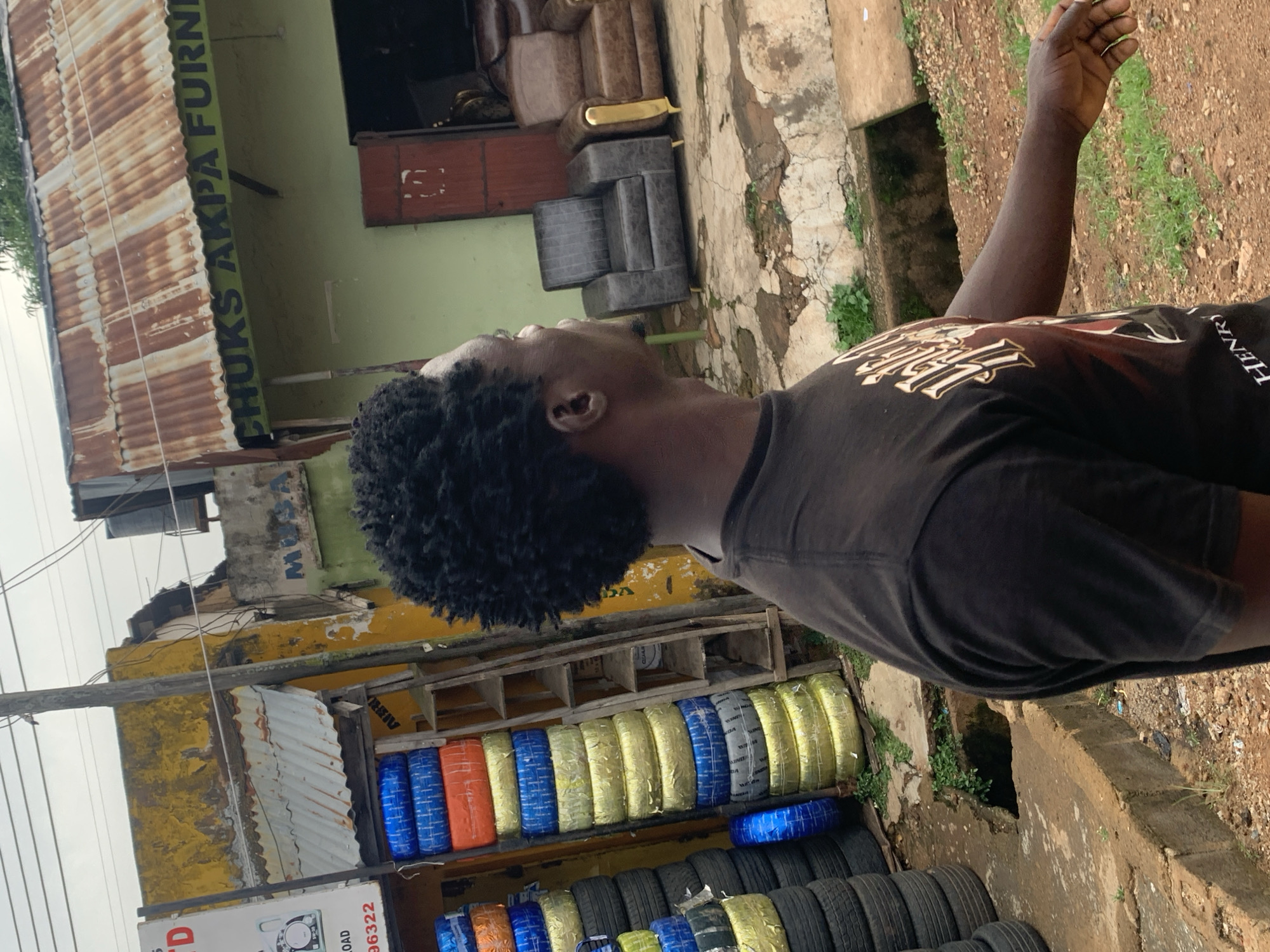 The image depicts a street scene in Jos, Nigeria. The foreground features a young Black person with dark, curly hair, wearing a black t-shirt with a graphic design. They are looking upwards and to the right, with their right arm extended upwards. The person appears to be standing outdoors on a paved surface, next to a roadside stall or shop.
Behind the person, a building with a green wall and a corrugated metal roof is visible. There is a sign on the building that reads "CHUKS AKPA FURNI". Further down the building, another sign reads "MUSA". Inside the building, furniture, including sofas and chairs, can be seen.
In front of the building, stacked on shelves, are rows of tires. Some of the tires are wrapped in brightly colored plastic, including blue, orange, and yellow. There are also piles of unwrapped black tires. Power lines are visible above the scene. The ground is a mix of dirt and concrete, with some grass growing in patches. The lighting suggests it is daytime, possibly overcast given the muted sky. The overall impression is of a busy commercial area in an urban or semi-urban setting in Nigeria.