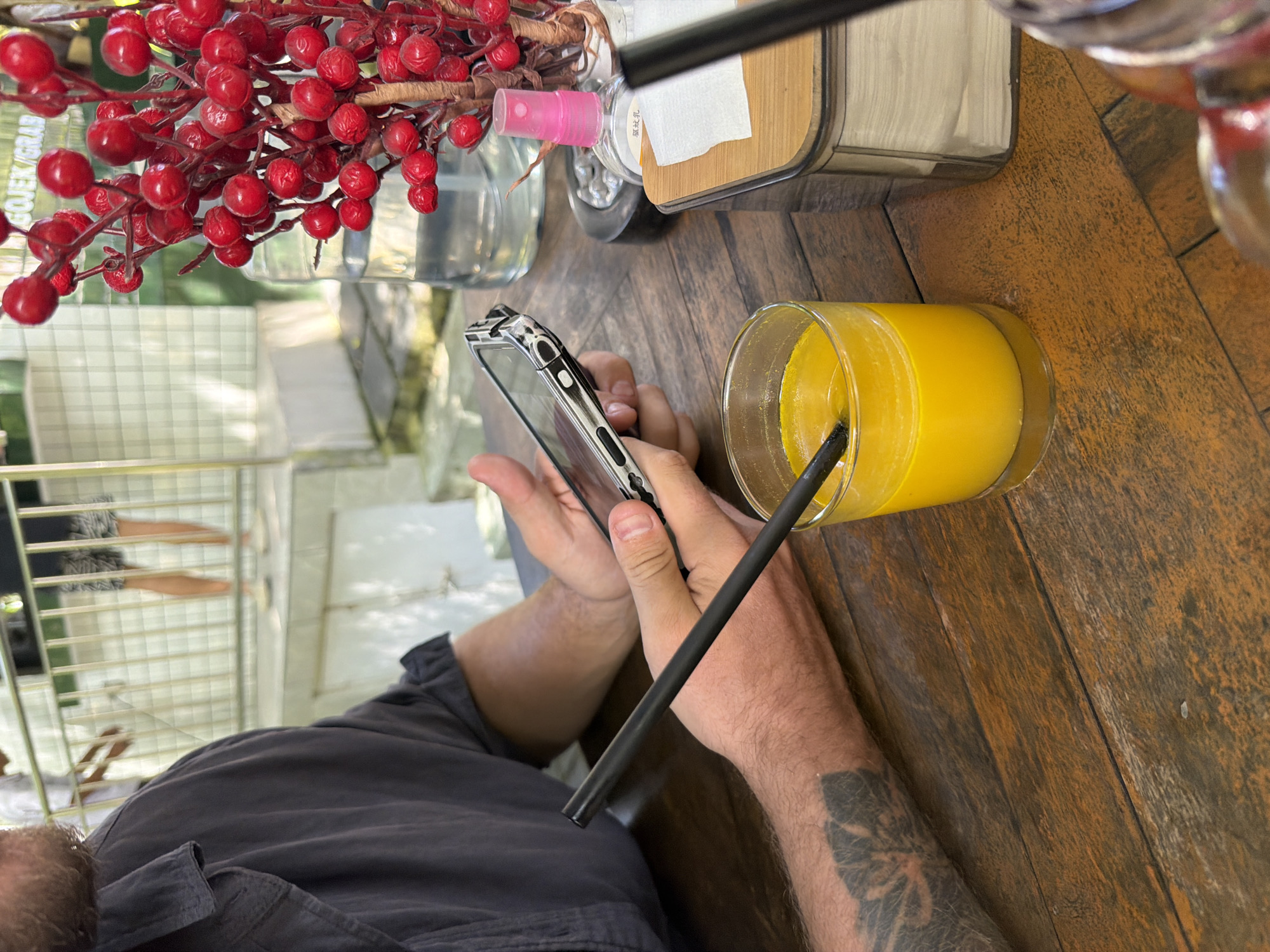 The image depicts a close-up view of a person sitting at a wooden table, holding a smartphone. The person's left arm, with a tattoo, rests on the table. In front of the person is a glass of orange juice with a black straw. To the right, a decorative branch with red berries is visible, along with a glass bottle of spray and a dispenser with white napkins. In the background, there's a blurred view of people standing behind a railing, suggesting an outdoor or semi-outdoor setting. The overall scene appears to be at a cafe or restaurant in Kuta Selatan, Indonesia, possibly near a public area given the railing and the presence of other people.