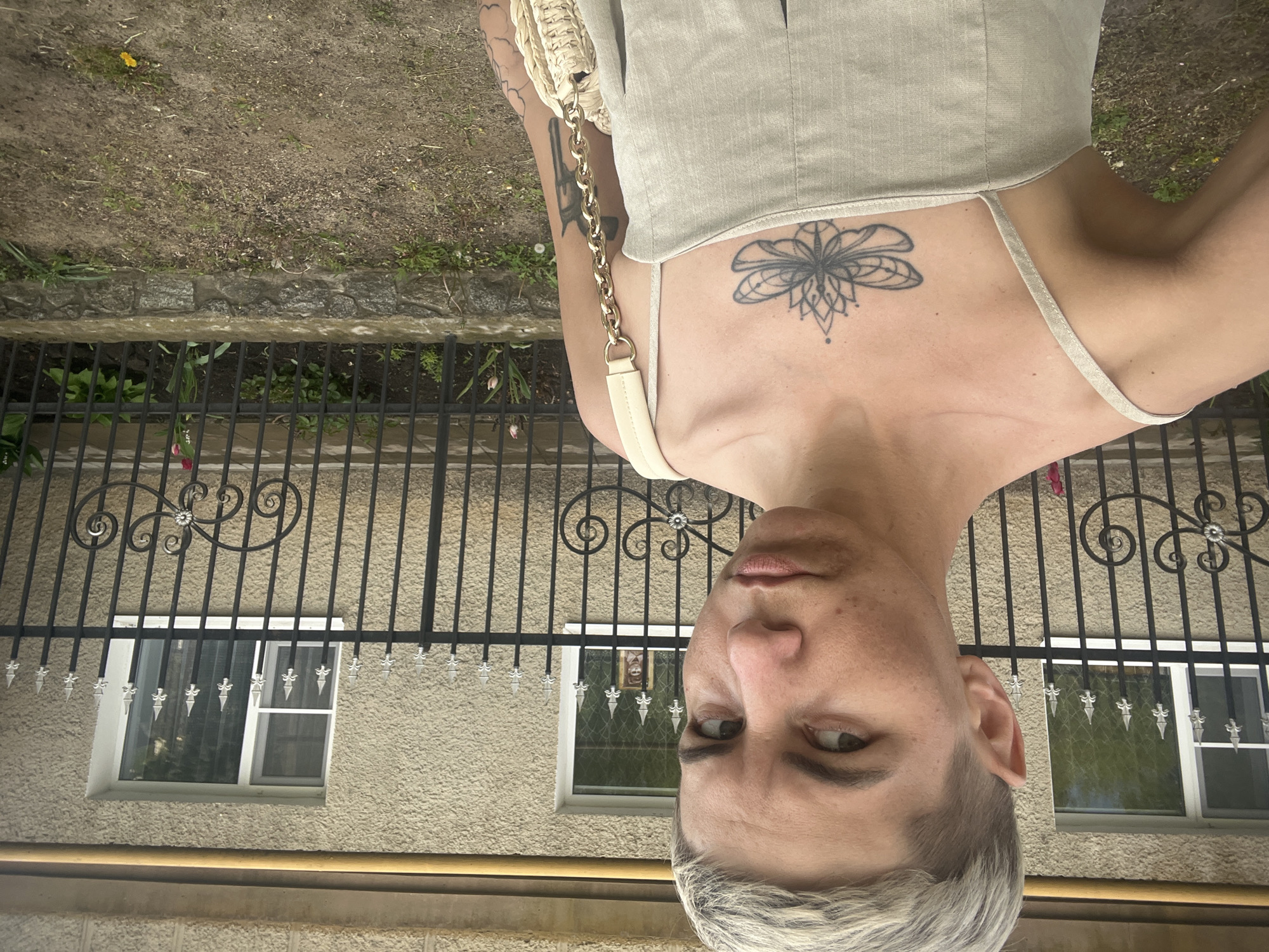 A person stands facing the camera with their head tilted to the side, framed by a wrought iron fence in the background, which suggests an outdoor urban setting. The fence is in front of a building. The person has short, light hair, and a chest tattoo. They wear a light-colored top with a thin strap. Part of a bag with a gold chain is visible on the left side. The location is Boguchar, Russia based on the building’s architecture and style of the fence.