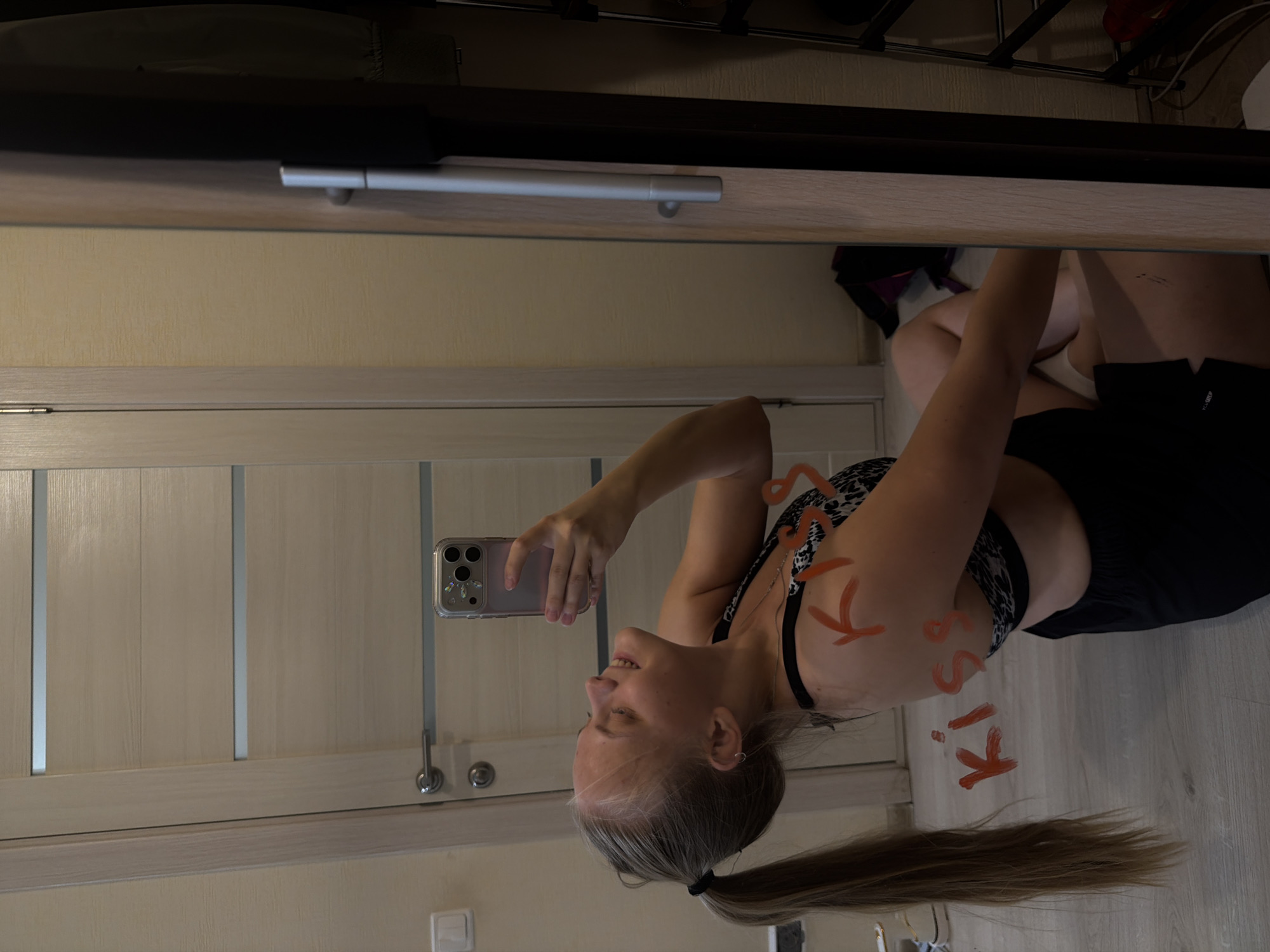 A young woman with blonde hair in a ponytail is sitting on the floor and taking a selfie in a mirror. She is wearing a sports bra and black shorts. The word "kiss" is written in red on her arm and shoulder. The background includes a door, a closet, and a shoe rack. The scene appears to be indoors, likely in a residential setting in Izhevsk, Russia.