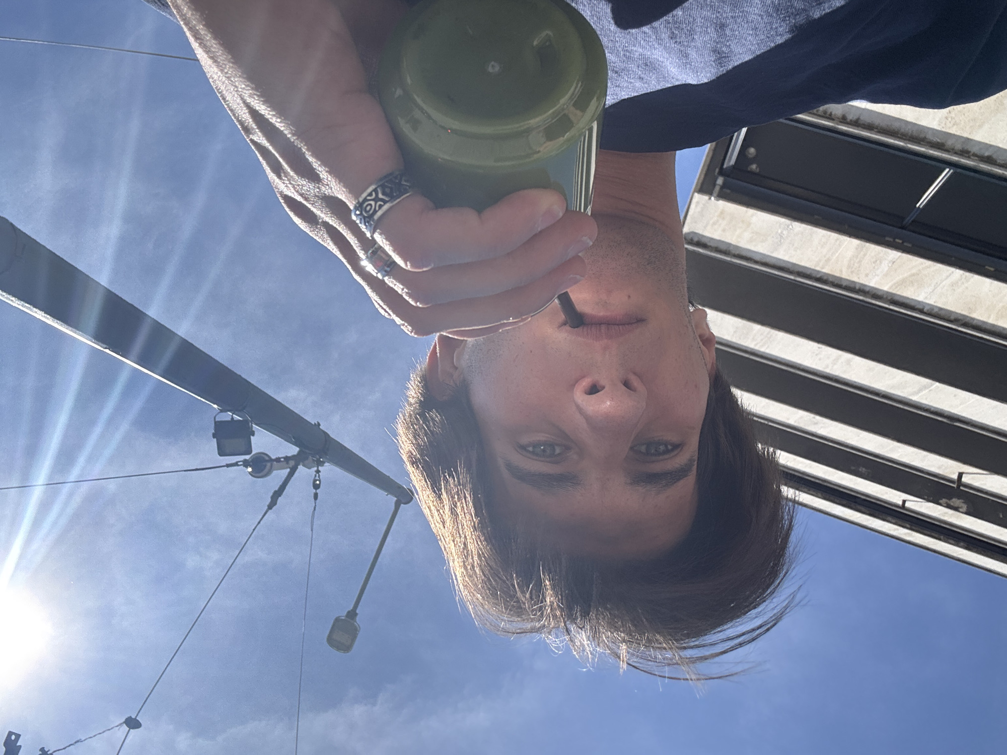The image is an upside-down selfie of a person holding a green beverage in a container with a straw. The person has short brown hair and blue eyes, and is wearing a ring on their hand. Behind the person is a blue sky and some architectural structures, including a pole with lights and a striped awning. While there is no specific information that would allow me to know its location, the city of Sofia is provided, so the scene is located in Sofia, Bulgaria.
