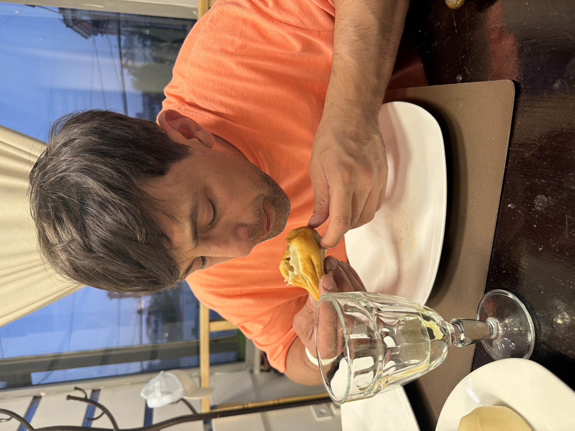 This image captures a man in an indoor dining setting, likely a restaurant or cafe in Krasnoyarsk, Russia. The main subject is a man with short dark hair and light stubble, wearing a bright orange t-shirt. He is shown from the chest up, leaning forward and looking down at a golden-brown, pastry-like food item, possibly a khachapuri, which he holds carefully in both hands. His expression is one of focused attention, suggesting he is either examining or about to take a bite of the food.
The table in front of him is dark brown, with a matching dark brown placemat. On the placemat sits a white, empty plate, suggesting he is either finishing or preparing to eat. A clear, textured stemmed glass, possibly a wine or water glass, filled with a clear liquid, is positioned to his right. Partially visible at the bottom right is another white plate holding a yellow condiment, possibly butter.
In the background, a large window offers a view of a clear blue sky and reflections of buildings, indicating it is daytime, possibly late afternoon. A light-colored curtain hangs to the left, and a white baseball cap rests on a structure further back, adding to the casual atmosphere of the scene. No visible text is present in the image.