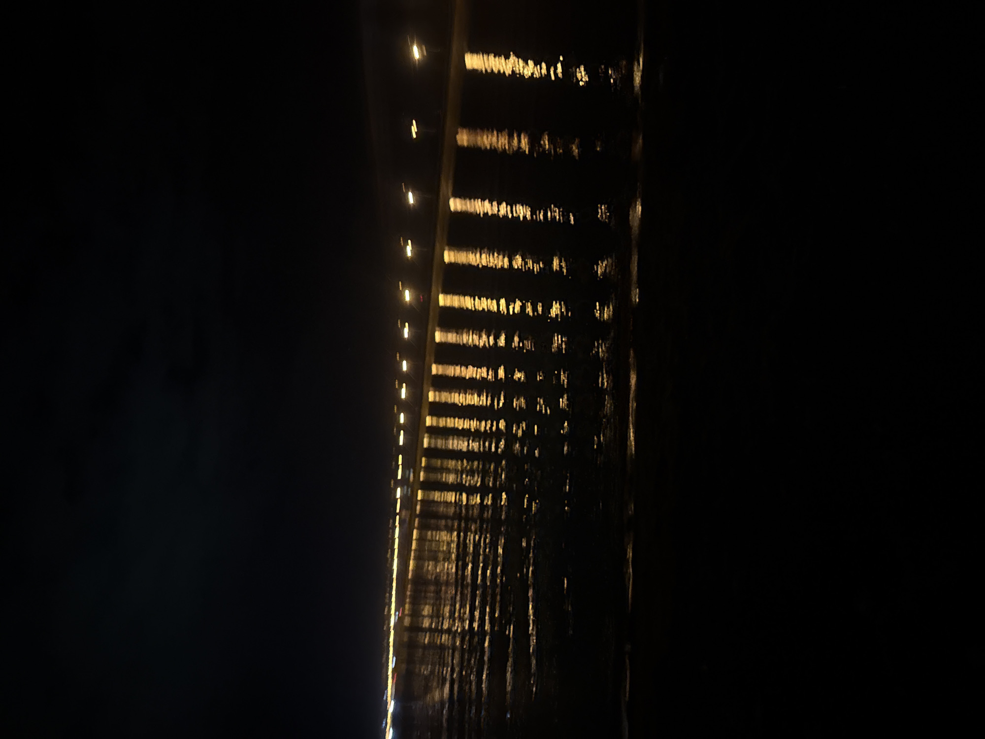 The image is a nighttime scene in Noviy, Russia, capturing a dimly lit landscape. The foreground shows a dark, wet surface, possibly sand or shallow water, with gentle ripples. In the midground, a row of streetlights casts elongated, shimmering reflections onto the water's surface. These reflections are a bright yellow-orange hue and appear as vertical streaks of light. Beyond the streetlights, a faint line of distant, blurred lights suggests buildings or a more developed area. The background is a dark, starless sky, with a subtle hint of clouds near the horizon. The overall atmosphere is dark and serene, with the artificial lights providing the only significant illumination. There are no people or animals visible in the frame.