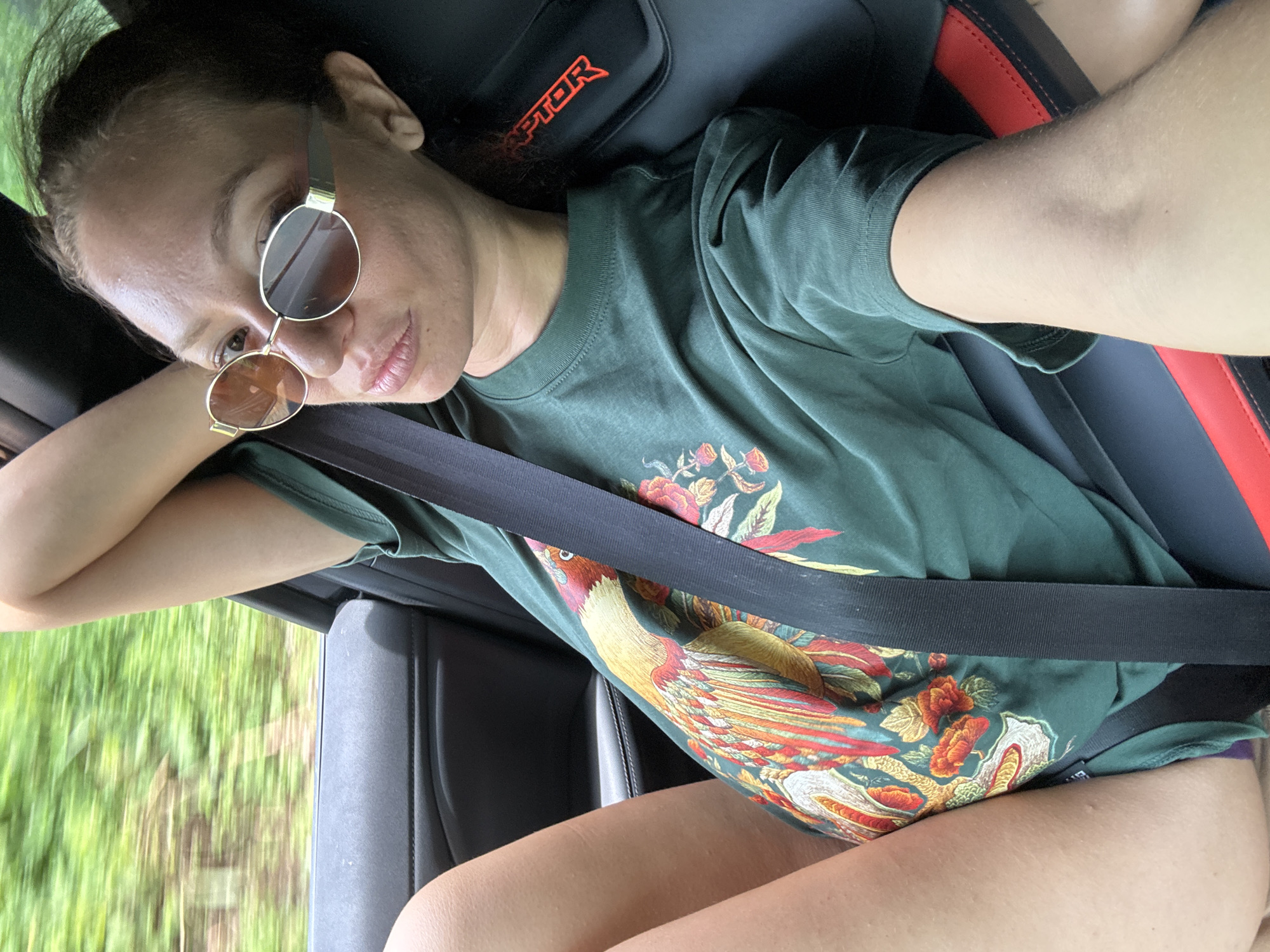 The image shows a young adult female seated in the passenger seat of a car, captured from a high-angle perspective. She has dark hair pulled back and wears stylish round sunglasses with gold frames, which partially obscure her eyes. Her expression is relaxed, with a slight pout. She is wearing a dark green t-shirt featuring a large, colorful graphic print of a bird, resembling a phoenix or pheasant, surrounded by elaborate floral designs. A black seatbelt is fastened across her chest and lap.
The car's interior is visible, including a black leather seat with red accents and a headrest that prominently displays the word "RAPTOR" in red text, suggesting the vehicle is a Ford Raptor or a similar model. Her right arm is bent, resting near the car's window frame. Outside the window, a blurred green landscape indicates the vehicle is in motion, likely traveling through a rural or natural area. The scene is brightly lit, suggesting it is daytime. The image captures a casual, relaxed moment during a car journey in Bảo Hà, Vietnam.