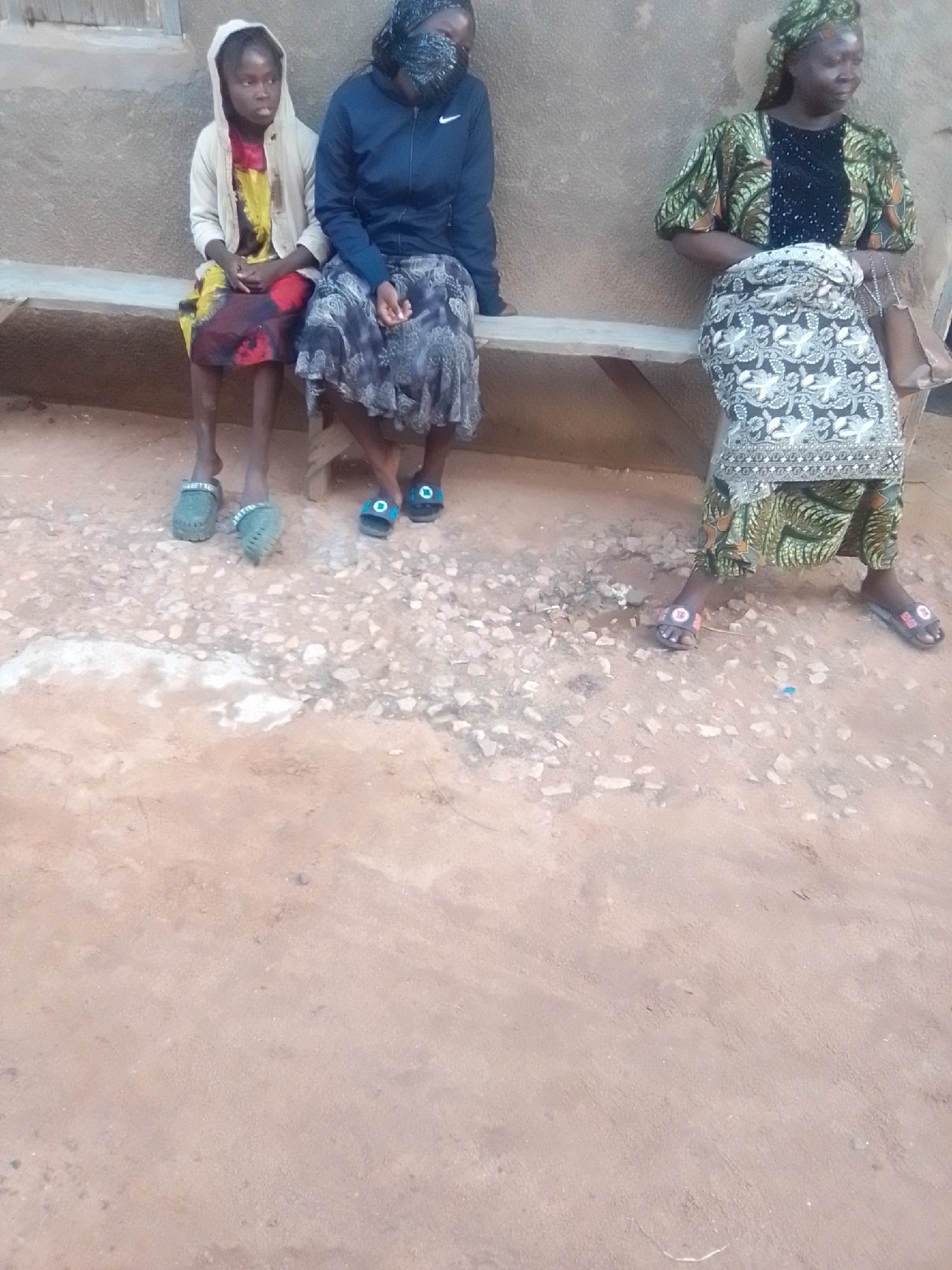 Three individuals are seated on a wooden bench against a rough-textured wall. On the left, a young person wearing a light-colored hooded jacket over a colorful dress sits with their feet in green clogs. To their right, another person is seated, wearing a dark blue jacket and a patterned skirt, with their face partially obscured by a patterned cloth covering their head and nose. On the far right, a third person, also wearing a headscarf, is dressed in a patterned green and black top and a patterned skirt, with their feet in sandals. The ground in front of the bench is a mix of reddish-brown dirt and small stones. The scene appears to be outdoors in a rural or semi-rural setting.