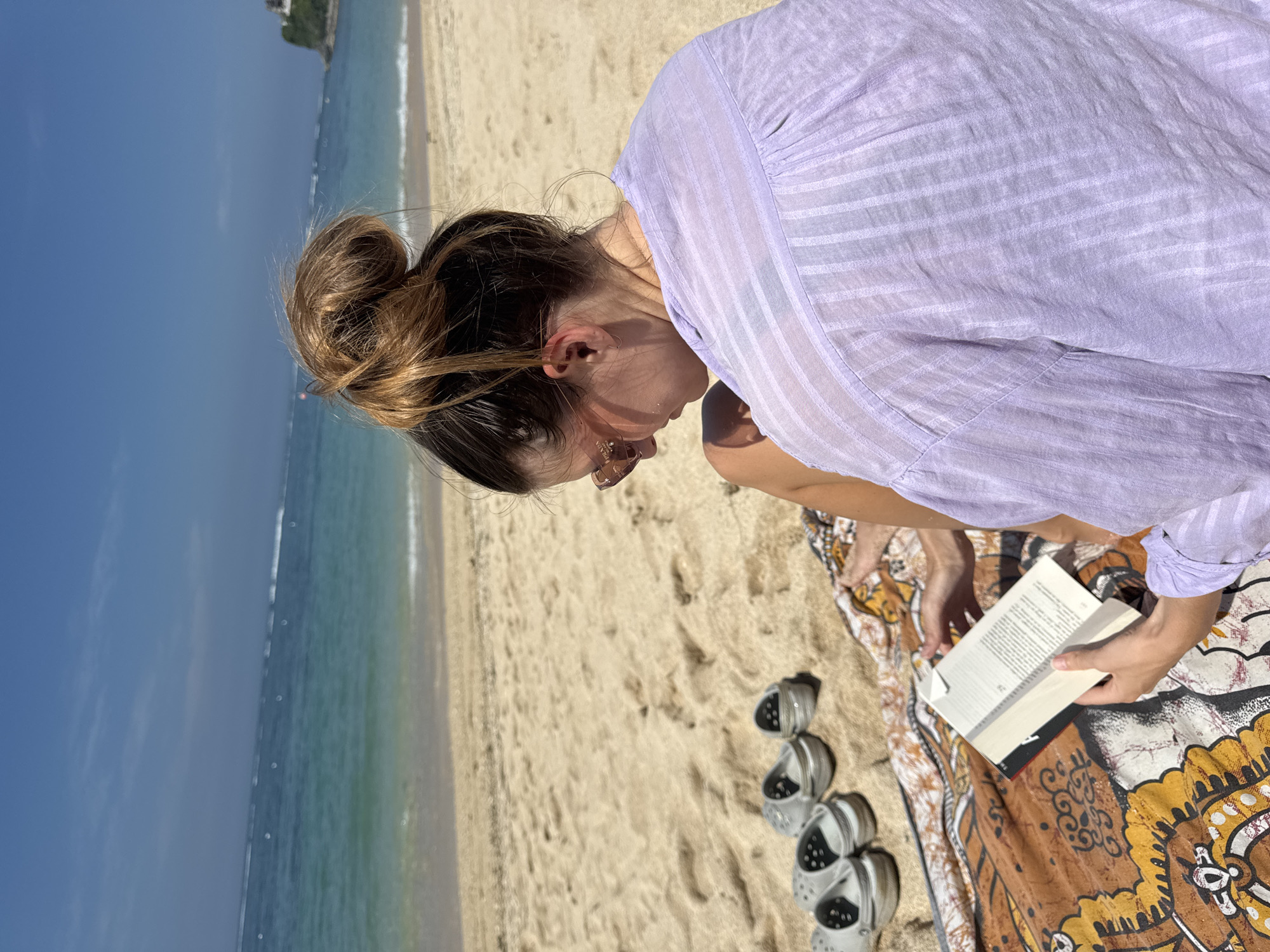 A woman with her hair in a bun is reading a book on a beach. She is wearing a light purple striped shirt and sunglasses. She is sitting on a patterned brown and yellow cloth, with a pair of grey Crocs next to it. The beach has tan sand, and the ocean is visible in the background under a blue sky. A small island or land mass can be seen in the far distance. The location is Unknown, Unknown.