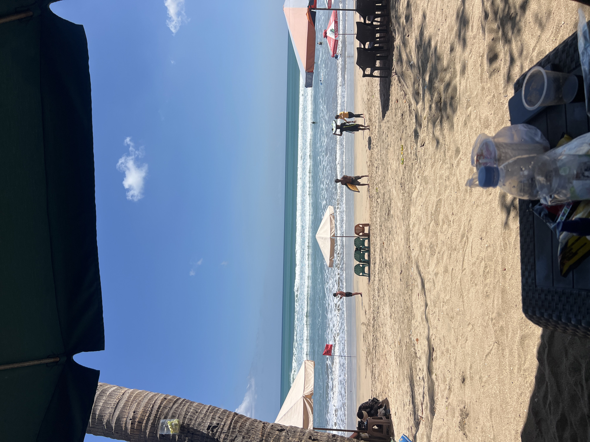 This is a medium shot taken from a seated perspective, looking out at Kuta Beach in Kuta, Indonesia. The foreground is dominated by a table with a wicker-like texture, on which rests a clear plastic water bottle, two plastic bags with items inside, and a clear plastic cup. The sandy beach stretches into the midground, dotted with beach chairs and umbrellas. A large palm tree trunk stands prominently on the left side of the frame. In the distance, the turquoise ocean meets the horizon under a bright blue sky with a few wispy clouds. Several people are visible on the beach, some carrying surfboards, and others walking or standing near the water's edge. The overall scene depicts a sunny day at a popular tourist beach.