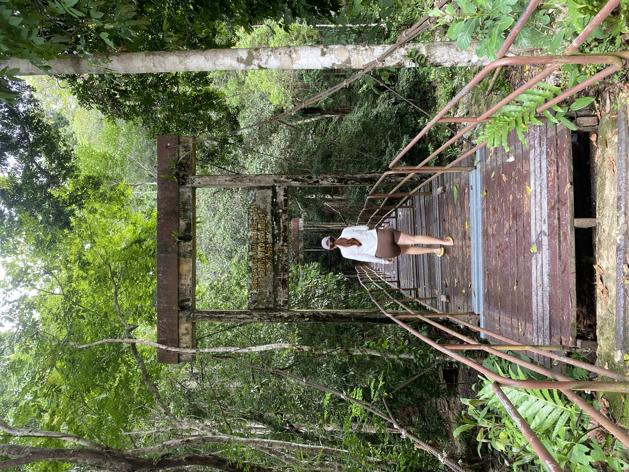 A medium shot captures a woman standing on a wooden suspension bridge within the lush greenery of Tambon Thai Mueang, Thailand. The bridge is constructed with brown wooden planks, bordered by rusty metal railings. The woman, positioned slightly off-center, is dressed in a light-colored jacket and shorts, complemented by yellow sandals. Above the bridge's entrance, a weathered sign displays text in Thai. The surrounding environment is densely vegetated with trees and foliage, casting diffused light throughout the scene. The camera angle offers a perspective looking down the bridge, enhancing the sense of depth within the image.