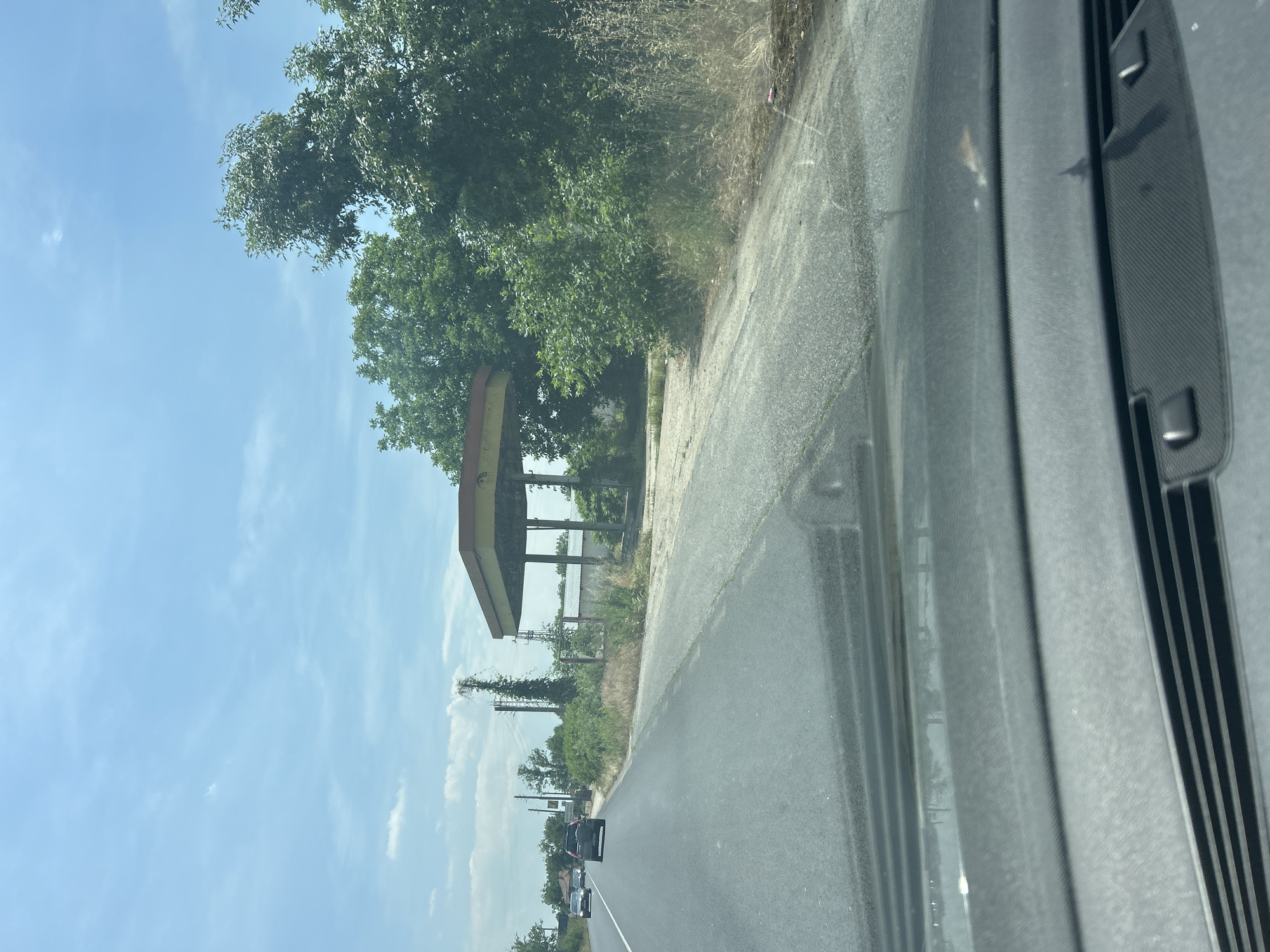 The image captures a roadside scene in Dolna Mahala, Bulgaria, viewed from inside a vehicle. On the left side of an asphalt road stands an abandoned structure, strongly resembling a former gas station canopy, characterized by its support pillars and a discolored, flat roof. The area around the structure and along the road is heavily overgrown with green trees and dense bushes, suggesting a prolonged period of disuse and neglect.
The road itself is a two-lane asphalt surface. In the distance, other vehicles are visible on the road, indicating ongoing traffic. The sky above is clear blue with scattered white clouds, suggesting bright, sunny weather, likely midday or early afternoon. Faint utility poles or power lines are discernible amidst the vegetation in the mid-ground.
The right edge of the main image shows a portion of a car's interior, likely a dashboard or windshield pillar, confirming the perspective from inside a moving or parked vehicle. No visible text or signage can be identified on the abandoned structure or within the frame. The scene evokes a sense of abandonment and nature reclaiming a once-functional roadside stop.