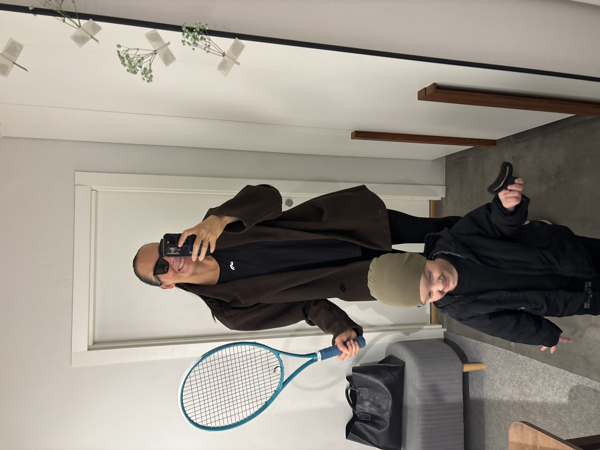 A mirror selfie features a person holding a blue tennis racket and a mobile phone, with a child standing to their right. The person in the mirror is wearing sunglasses and a brown coat over a black t-shirt. The child is wearing a black jacket and a light green beanie. A gray bench and a black bag are positioned below the person holding the racket. Overhead, the ceiling is decorated with small bunches of flowers taped to the surface. There is no information to confirm if this scene took place in Chaikovsky, Russia.