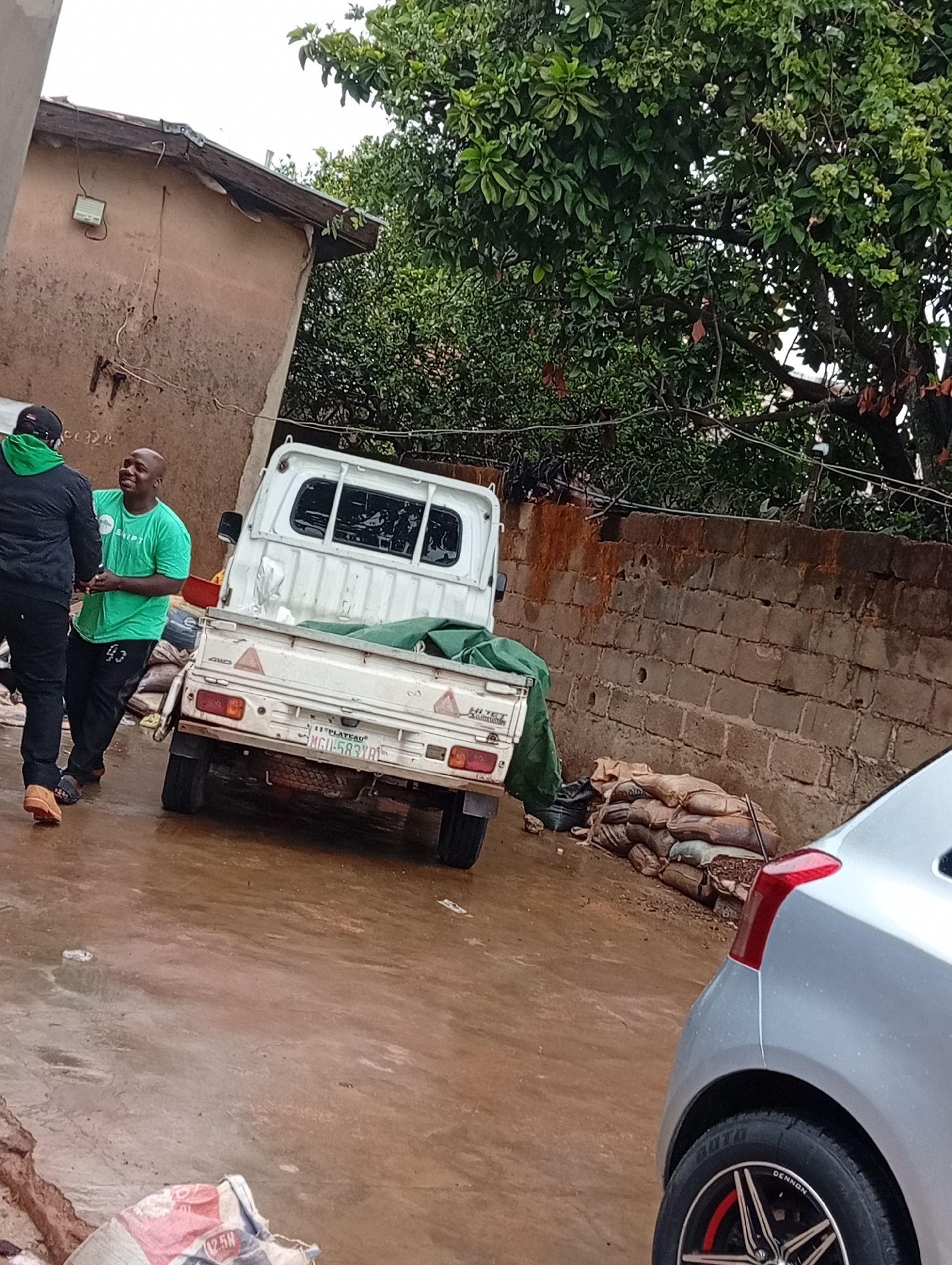 The image captures an outdoor scene in what appears to be a residential area. Two men are interacting near a small white truck, possibly a Kei truck, which is parked on a wet or muddy surface. The man closest to the camera is wearing a green t-shirt with the word "SHIPT" visible, black track pants with the number "93" on them, and brown sandals. The other man is wearing a black jacket with a green hood, black pants, and brown shoes.
The truck has a license plate that reads "MGU-583 YA" and the word "PLATEAU" is also visible. Behind the truck, along a brick wall, are several bags of what appears to be cement or other construction materials. A silver car is partially visible in the foreground on the right side of the image, showing the rear tail light and a portion of the tire and rim.
The environment includes a building with a brown exterior to the left and a tree with dense green foliage in the background. The general atmosphere suggests a rainy day. The location is specified as Gura Top, Nigeria.
