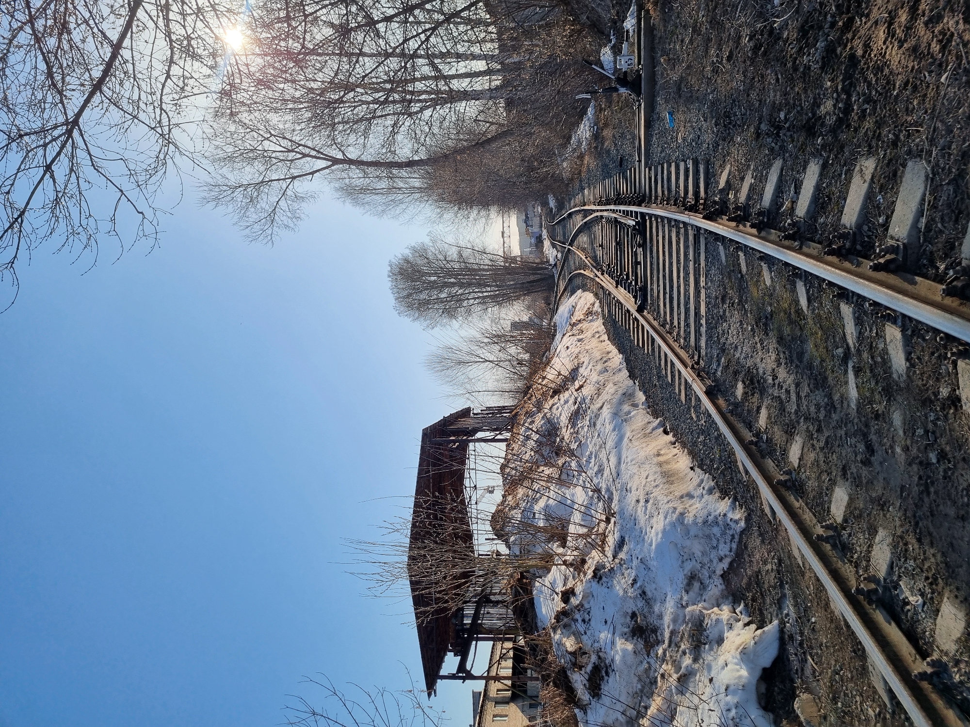 The image shows a section of railway tracks curving to the right. The tracks are set on concrete sleepers and run along a dirt and gravel bed. Patches of snow are visible on the ground beside the tracks. In the background, there is a dilapidated wooden structure with a partial roof, indicating possible abandonment or disrepair. Bare trees are present both above and to the side of the tracks. The sky is a clear blue with the sun visible at the top of the frame. The location is Chaikovsky, Russia.