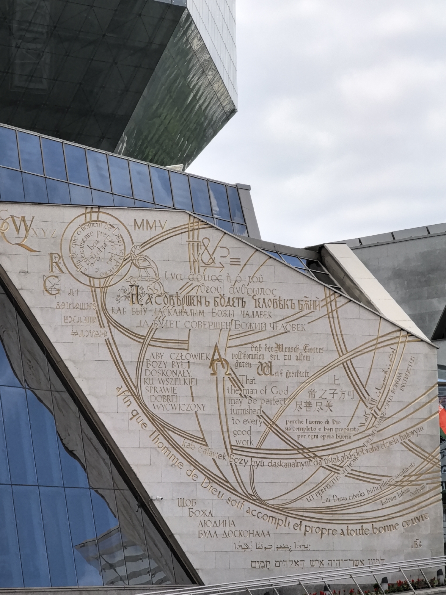 The image shows a large, light-colored stone wall adorned with a golden, intricate design featuring text in various languages and scripts. The wall is part of a modern building with large glass windows, reflecting the sky and other structures. The design on the wall appears to be a religious text or affirmation, with circular motifs and flowing lines connecting the different sections of text. Some of the scripts visible include Latin, Cyrillic, Greek, Arabic, Hebrew, and what appear to be Chinese characters. The overall impression is one of a monumental artwork intended to convey a message of universal significance. The environment suggests an urban setting, possibly a public square or the exterior of a significant institution.