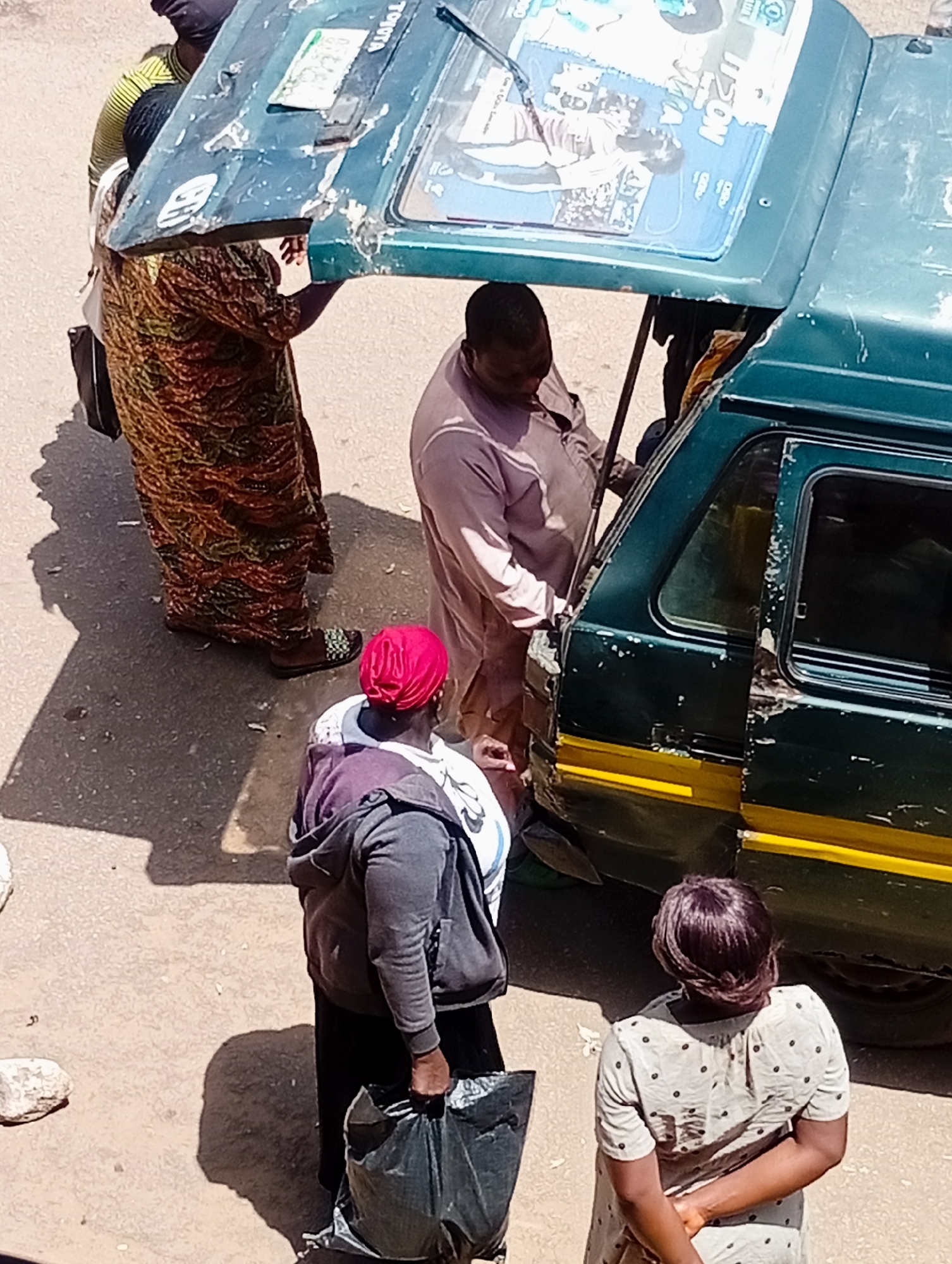 The overhead shot captures a scene on a dusty, paved street in Bukuru, Nigeria, where several people are interacting around a damaged green vehicle, possibly a public transport vehicle. A woman on the left, wearing a vibrantly patterned brown, orange, and green dress, carries a black bag over her left shoulder. She appears to be engaged with the vehicle. In the center, a man in a light pink garment is leaning into the vehicle, seemingly attending to something or speaking with someone inside. Another person, wearing a purple hooded jacket and a red headscarf, stands behind and to the left of the man, also appearing to interact with the vehicle, and holds a large black plastic bag. On the right side of the frame, a woman in a light-colored polka-dot dress stands with her back to the camera, her hands clasped behind her. The vehicle's rear section appears dented and scratched, with part of the roof lifted, revealing some posters or images attached to the underside of the lifted section. A yellow stripe runs along the side of the vehicle. The ground is rough and uneven, with scattered stones. The lighting suggests it is daytime, with strong shadows cast on the ground.