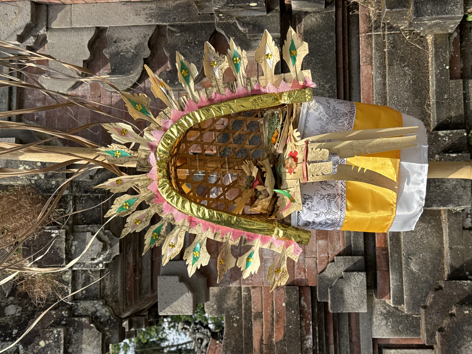 This image captures an intricately decorated ceremonial offering, likely a "Canang Sari" or similar Balinese devotional item, positioned in front of a stone temple structure. The offering is a vibrant display of natural materials, featuring a woven bamboo basket at its apex, adorned with golden leaf-shaped decorations tipped with green and gold accents. Pink fabric trims the arching top of the basket. Beneath the basket, the offering is draped with white fabric printed with detailed black line drawings of figures, followed by a bright yellow silk fabric, and finally a white fabric base. Dried palm leaves and other natural elements extend upwards, forming a crown-like structure. The backdrop is a weathered, multi-tiered stone structure, typical of Balinese temple architecture, constructed with rough-hewn blocks and accented with red brickwork. The lighting suggests it is daytime, with shadows indicating the sun's position. The specific location in Gianyar, Indonesia, is implied by the cultural context of the offering and the architectural style. No people are present in the scene, and the focus is entirely on the religious artifact and its temple surroundings.