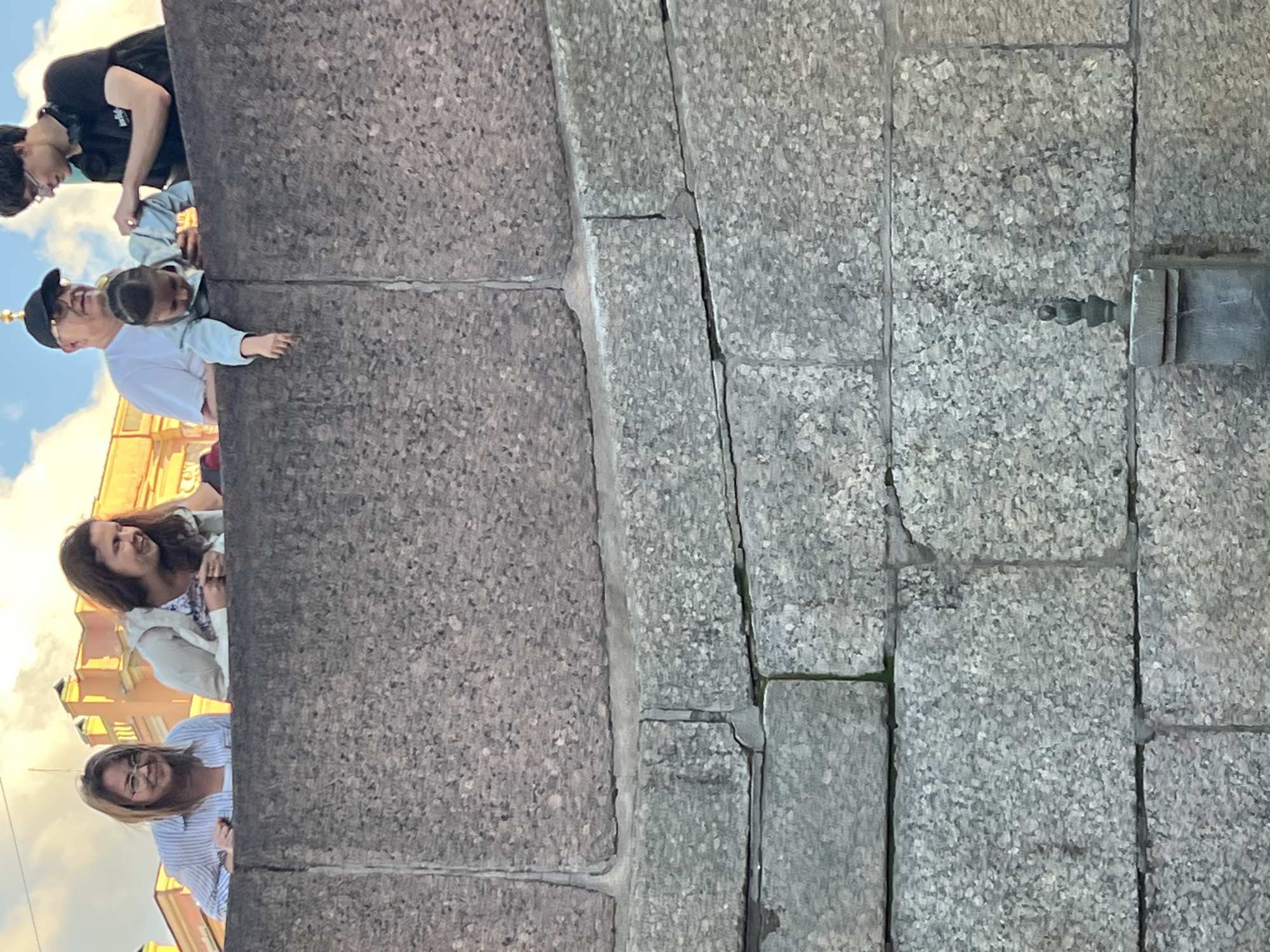 A group of people, including adults and a small child, are looking over a stone wall. The child is leaning over the wall, with their hand extended. The people appear to be on a promenade or elevated walkway. In the background, there are buildings with ornate architecture, characteristic of St. Petersburg, Russia. The sky is partly cloudy. Below the wall, the stonework continues, with some visible cracks. The scene suggests a casual gathering of a family or friends enjoying a view.