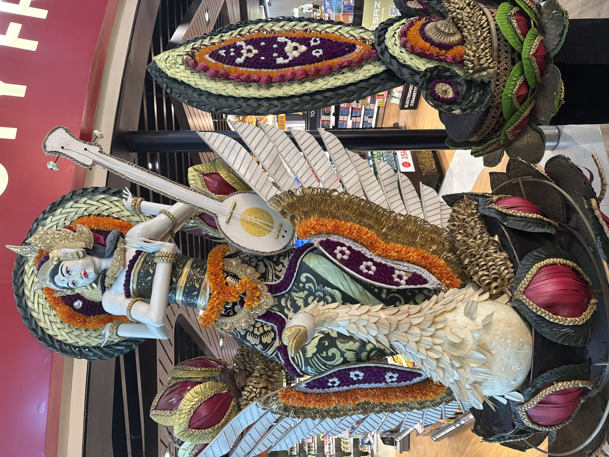 This is a medium-shot, eye-level, indoor photograph of a large, ornate Balinese sculpture, likely a representation of a Hindu deity, possibly Saraswati, given the musical instrument. The sculpture is richly decorated with woven materials, floral arrangements, and what appears to be painted wood or plaster. The deity has pale skin, dark hair styled with elaborate headwear, and is adorned in traditional clothing with intricate patterns and gold accents. She is holding a stringed instrument resembling a lute. The sculpture is supported by a base that incorporates elements resembling a swan or goose, along with large, wing-like structures and layered, petal-shaped decorations. The surrounding environment suggests a public space, possibly an airport or a shopping mall, with displays of merchandise and signage visible in the blurred background. The lighting is bright and even, illuminating the details of the sculpture. The image's location is identified as Kuta, Indonesia.