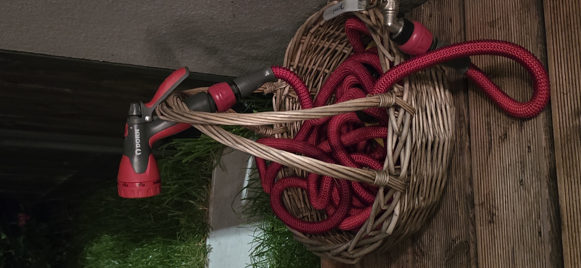 A red and gray Dorn brand hose nozzle is attached to a coiled red hose within a wicker basket. The basket has two handles made of twisted wood. The hose nozzle is black and red with a rotating collar for spray pattern selection, labeled "MIST," "FLAT," "JET," and "SHOWER." The nozzle also has a red trigger. The hose appears to be a fabric-covered expandable hose. The basket sits on a weathered wooden deck. In the background, there is green grass, a light-colored planter, and a dark vertical fence or wall. To the right of the basket, a silver faucet with a red connector is visible. The image appears to be taken at dusk or night, with artificial lighting. There are no people or animals present. The scene depicts gardening equipment stored outdoors. The specific location in Kaliningrad, Russia cannot be determined from the visual information provided.