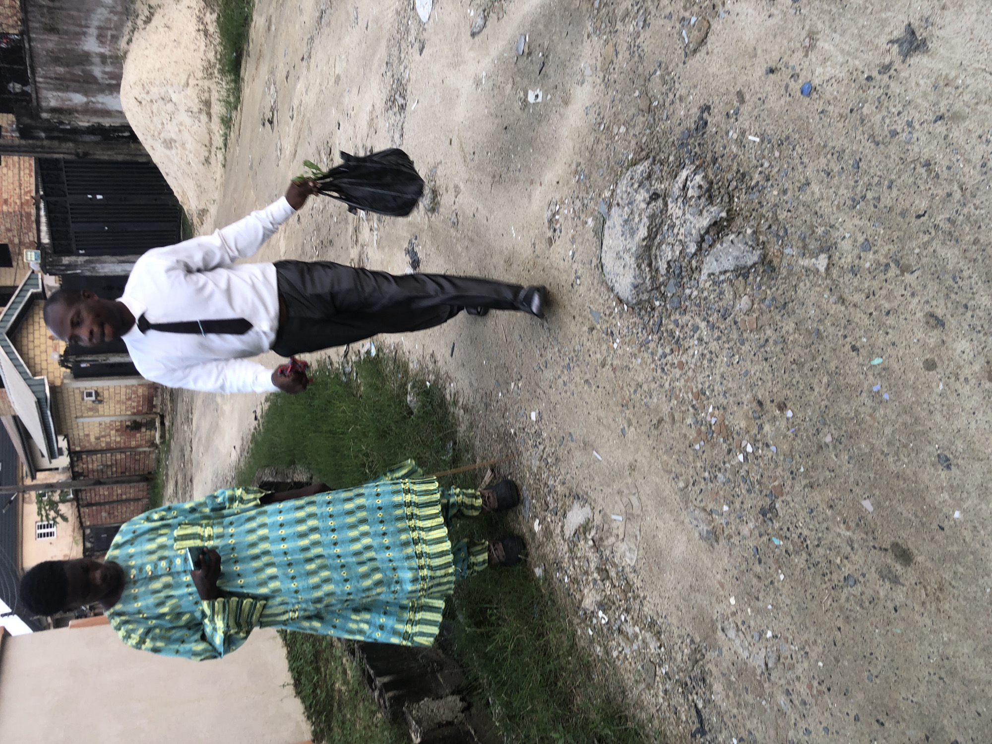In the city of Ado, Nigeria, two men walk along a dirt road. The man on the left wears a traditional blue and green patterned tunic and trousers, holding a mobile phone in his right hand. He also uses a walking stick. The man on the right is dressed in a white button-down shirt, a black tie, and dark trousers, carrying a black plastic bag in his left hand. The environment is characterized by a dirt road with scattered debris, grass on the sides, and buildings in the background. A large pile of sand is visible behind the man on the right, suggesting ongoing construction or landscaping. The sky appears overcast, casting a soft light on the scene.