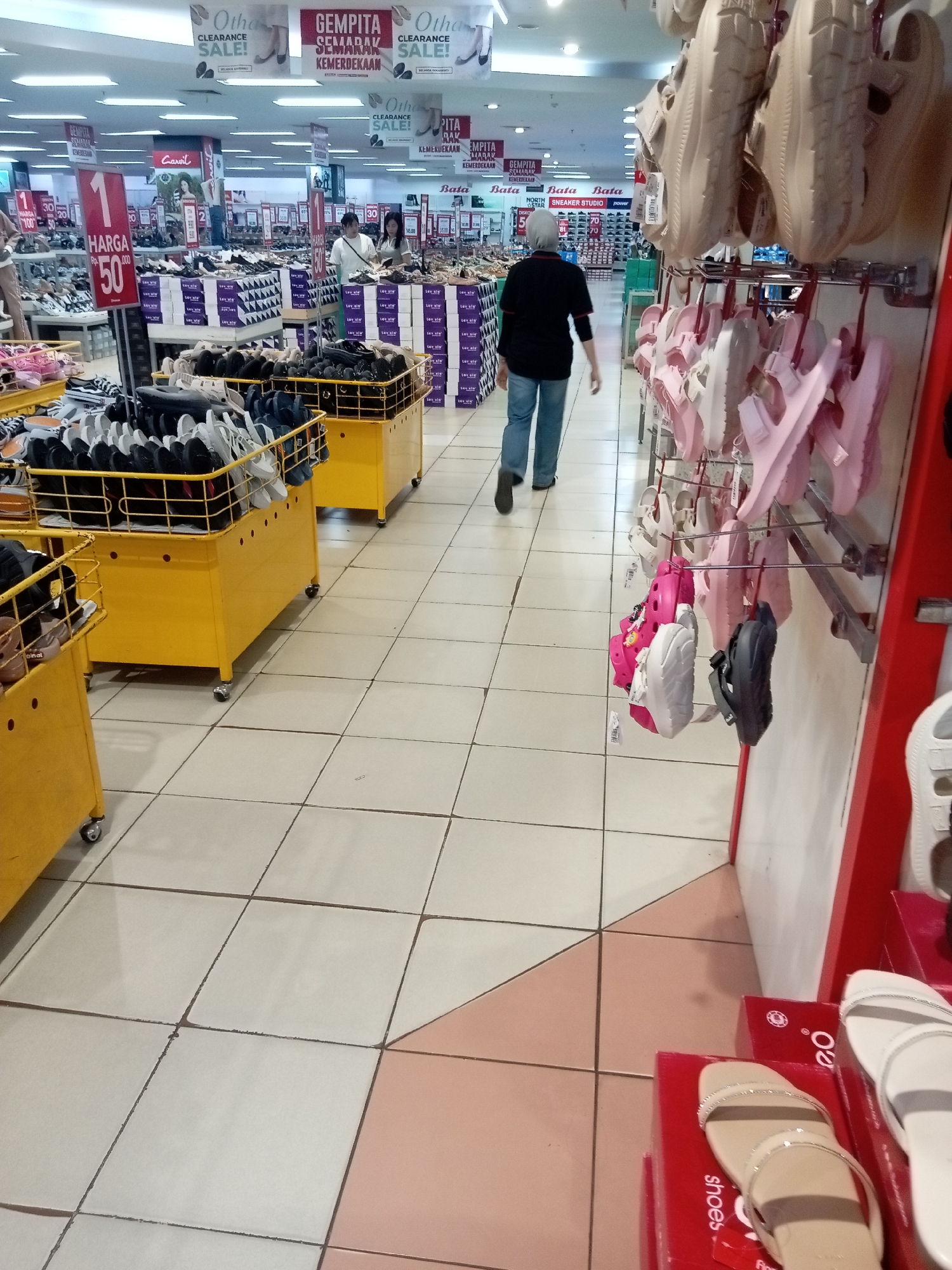 This image depicts the interior of a shoe store, likely a department store or a dedicated shoe retailer, in Batam, Indonesia. The environment is brightly lit with fluorescent overhead lighting and features rows of shelves and display stands filled with footwear. The floor is tiled with a light beige and pink pattern.
In the foreground, several yellow metal carts are filled with various types of shoes, including sandals and flip-flops, displayed in an organized but somewhat crowded manner. To the right, a display rack showcases an assortment of pink and white sandals. Further back in the store, more shoe displays and shelving units are visible, with some featuring promotional signage for "Clearance Sale" and "Gempita Semarak Kemerdekaan."
Two women are present in the middle ground of the image. One, wearing a white top and green pants, is looking at shoes on a display. The other, in a black shirt and blue jeans, is walking away from the camera, towards the back of the store. In the background, more shoppers and store staff can be seen browsing or working. The overall impression is a busy retail environment, typical of a large shopping mall or a busy street-level store in a commercial area of Batam.