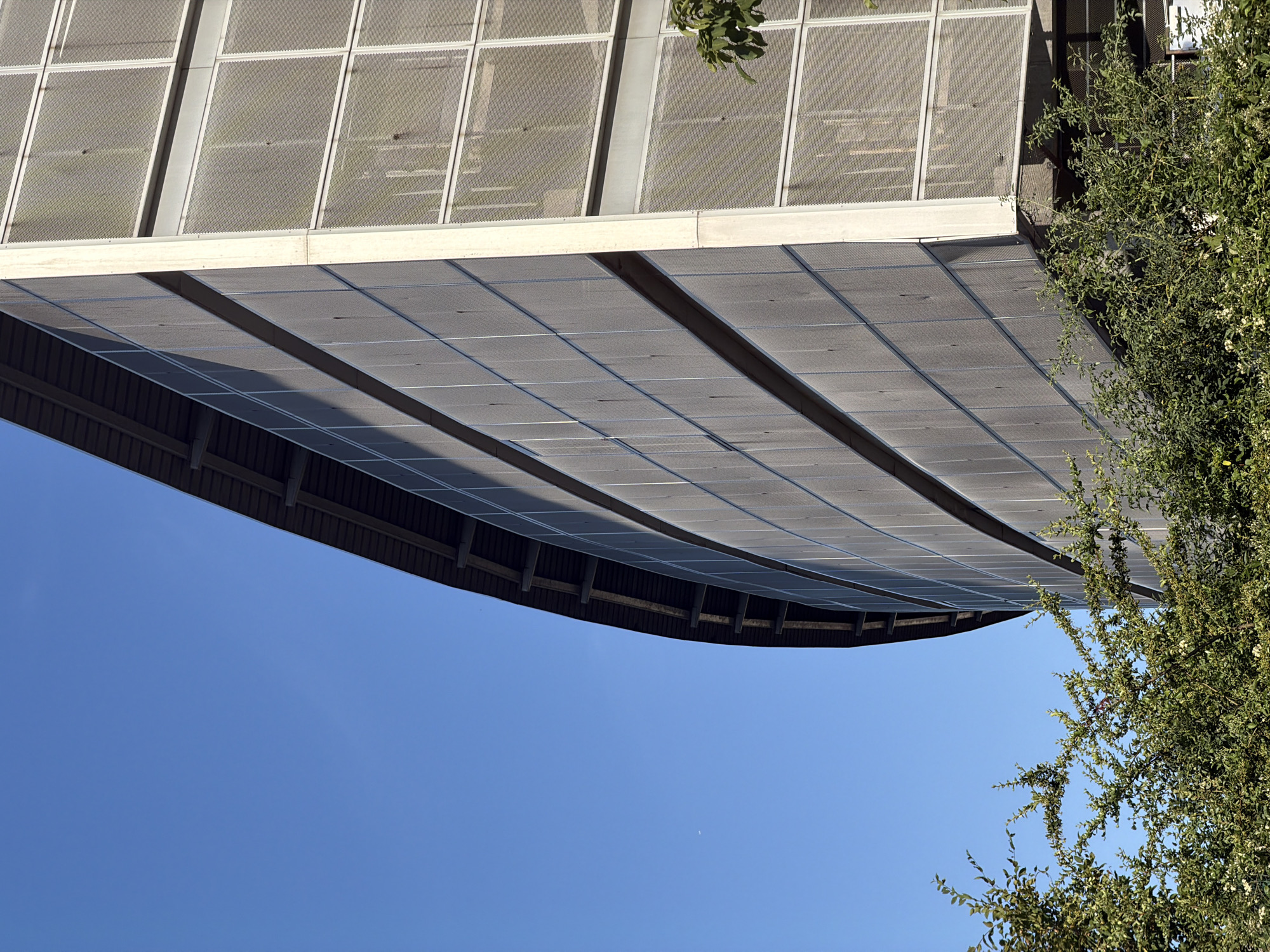 The image shows an upside-down view of a modern building with a distinctive facade. The building appears to be covered in solar panels on its lower section, which curves outwards. Above the solar panels, there are large, rectangular windows or panels with a grid-like pattern. A lush green tree with dense foliage and small white flowers is visible on the right side of the frame, partially obscuring the building's facade. The sky is a clear, bright blue, suggesting a sunny day. The perspective is unusual, as the building and the tree are seen from below, against the sky. There is no visible text in the image. The scene evokes a sense of urban architecture integrated with nature, with a focus on sustainable energy solutions. The bright sunlight casts distinct shadows on the solar panels, indicating it is likely daytime. The location is identified as Lyon, France, which is consistent with modern architectural styles and the presence of greenery in urban environments.