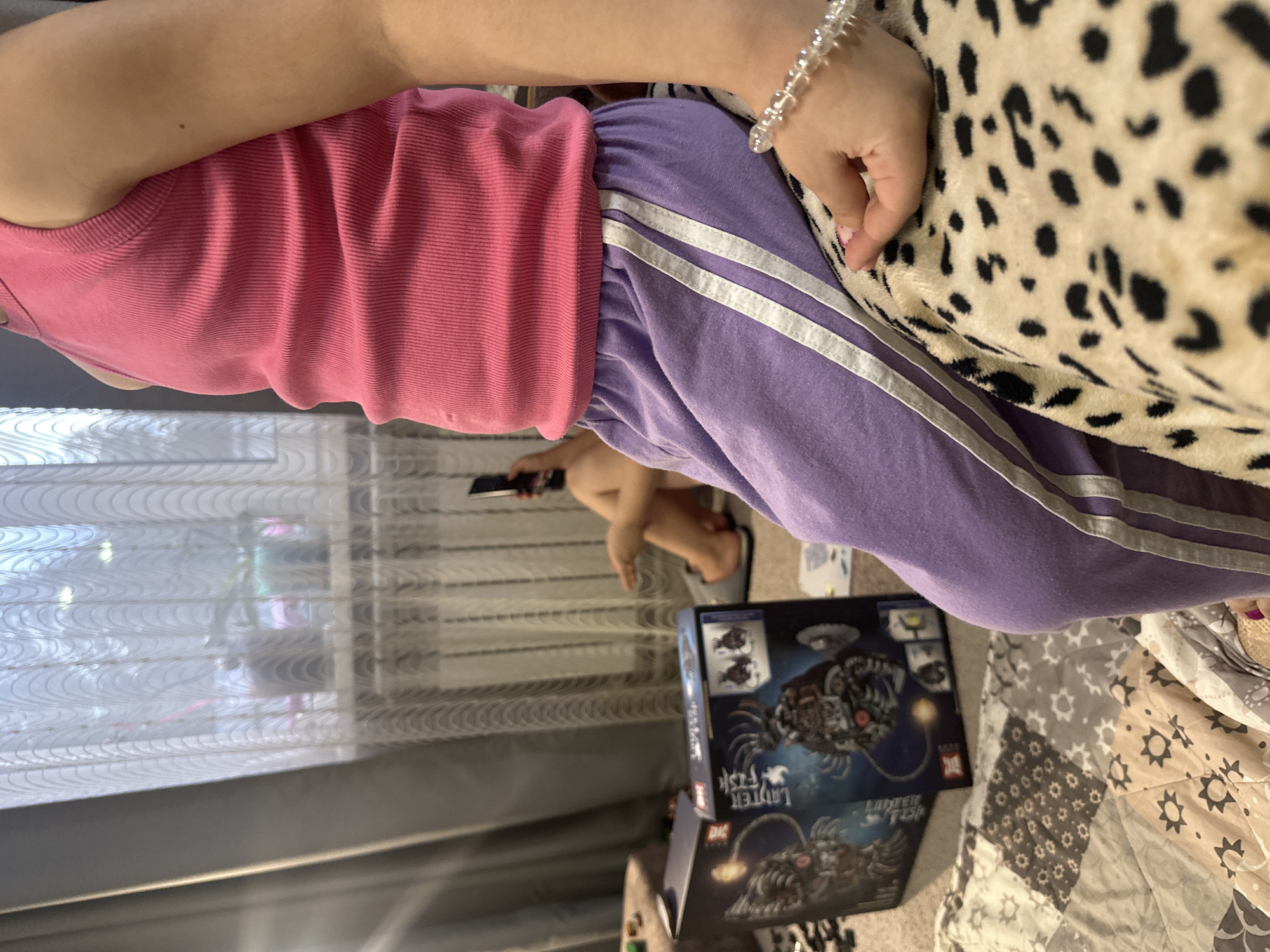 A person sits on a patterned bedspread near a window with sheer curtains. They wear a pink tank top and purple athletic pants with white stripes. Their hand, adorned with a beaded bracelet, rests on a leopard-print blanket. A box labeled "Lanтер FISH" is on the floor. The location is an indoor setting.