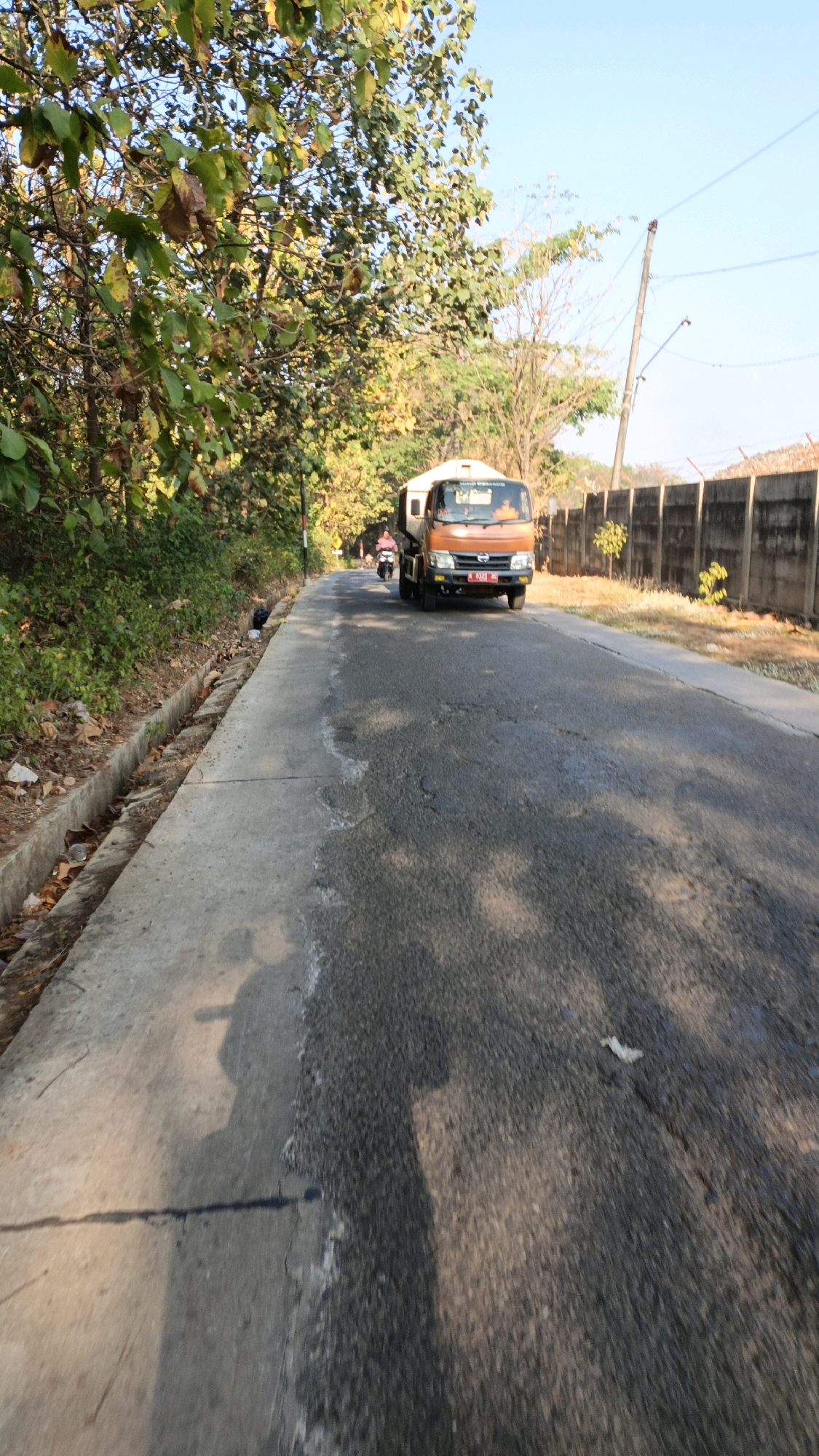 This is a ground-level, eye-level, wide shot of a paved road in Kuwasen, Indonesia. A bright orange truck is driving away from the camera, in the middle of the road. A person is riding a motorcycle behind the truck. The road is lined with green trees and bushes on the left, and a concrete fence on the right. The sky is clear and blue. There is a concrete drainage ditch on the left side of the road. The road surface has patches of wear and tear. The sunlight casts shadows on the road.
