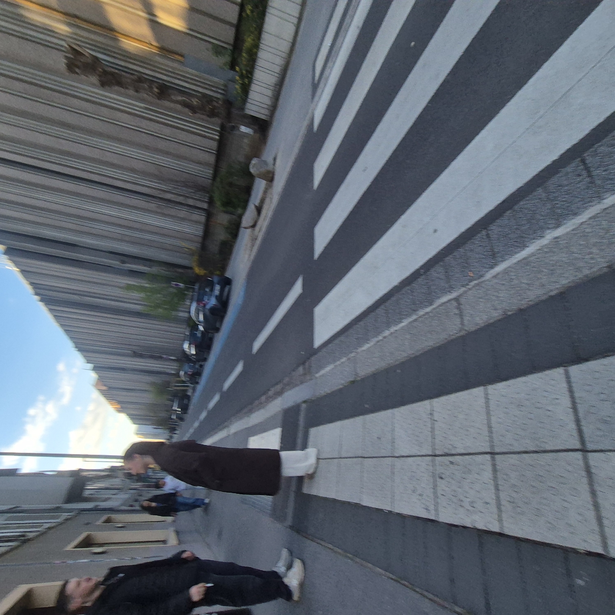 The image depicts an outdoor scene in Strasbourg, France, focusing on a street area with a crosswalk. A tall building with horizontal lines is visible on one side of the street. There are at least three people in the frame. One person is wearing a long brown coat with white footwear and is seemingly walking across the crosswalk. Another person is wearing a white shirt and jeans. A third person is lying on the ground, wearing black clothing, potentially holding a small item in their hand. Cars are parked along the street. The overall impression is of a typical urban environment.