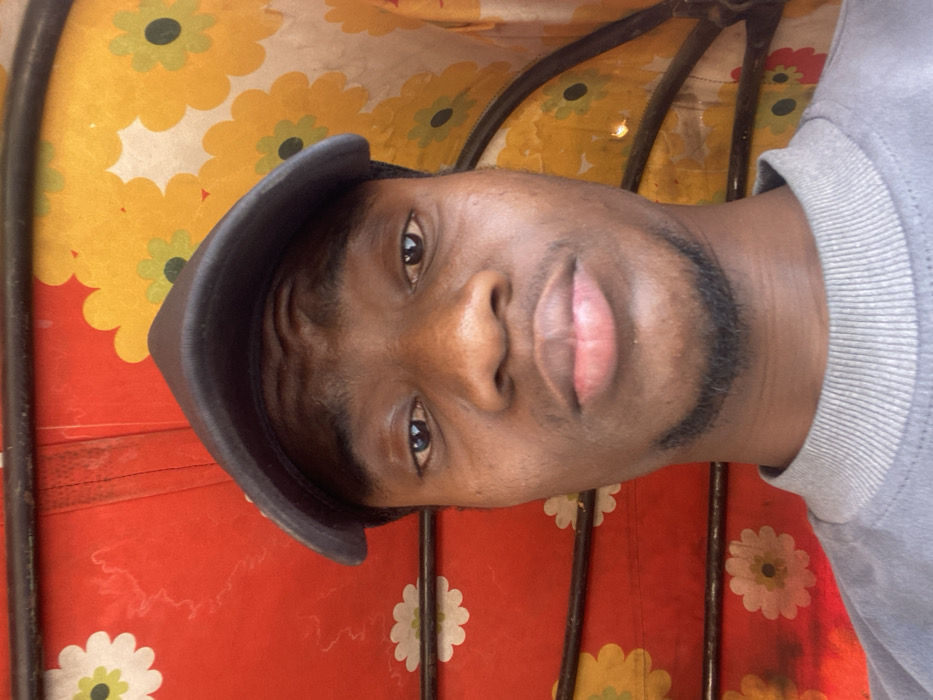 A young adult male, appearing of African descent, is depicted in a close-up, head-and-shoulders portrait shot, likely within the interior of a vehicle in Baganakwo, Nigeria. He wears a dark grey baseball cap and a light grey crew-neck sweatshirt with a ribbed collar. His gaze is directed forward towards the camera, maintaining a neutral expression. His facial features include dark skin, dark eyes, full lips, and short dark stubble along his jawline and chin. The background consists of a brightly patterned fabric, predominantly red on the left and yellow on the right. Both sections feature a repeated floral motif: white flowers with light green centers on the red background, and light green-centered flowers on the yellow background. Two horizontal dark metal bars traverse the background, indicative of structural components within the vehicle's canopy or frame. A vertical seam is visible on the red fabric to the left of the subject's head.