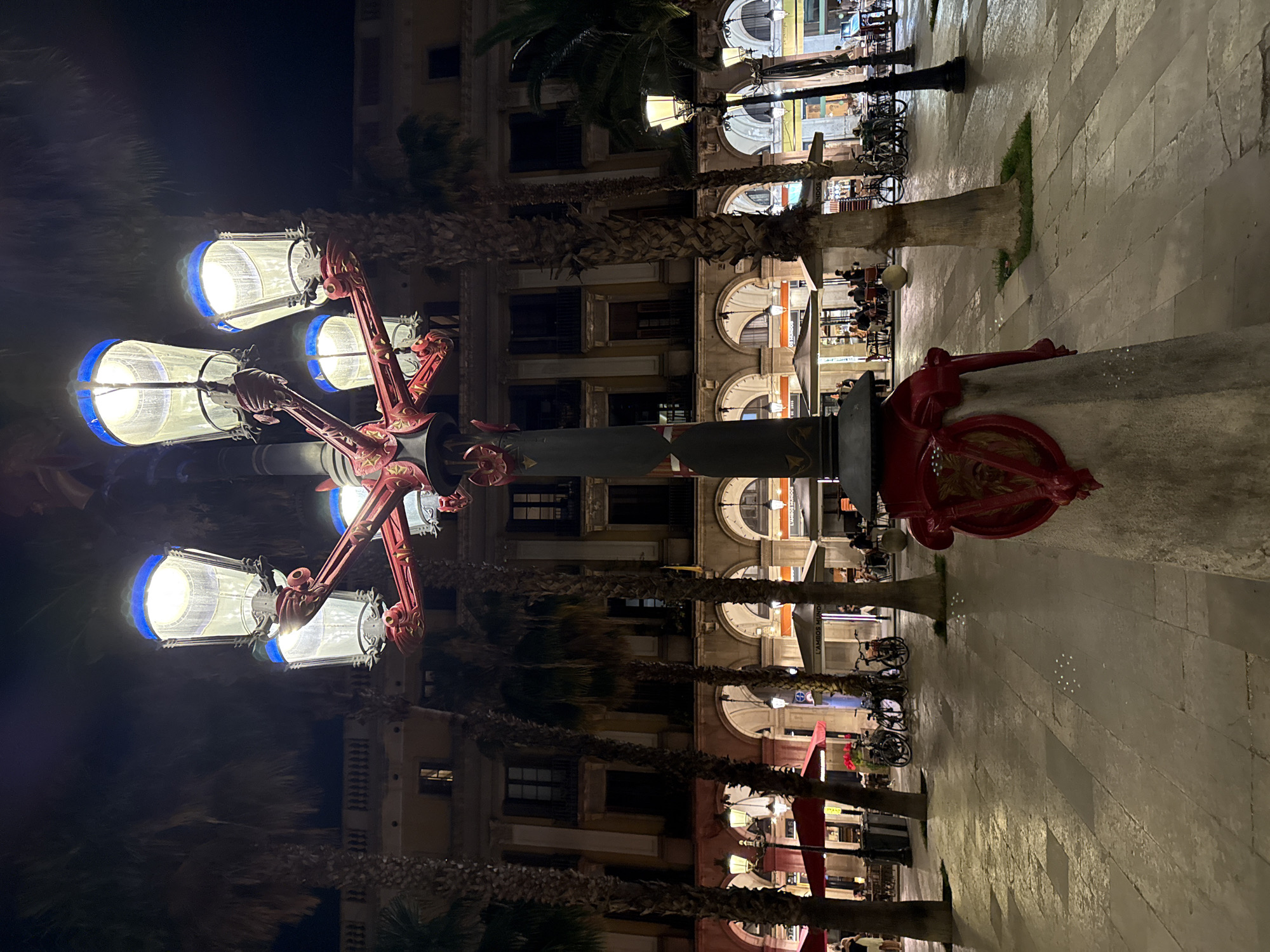 The image shows a street lamp in the foreground at night, with multiple lights illuminating the scene. Behind the lamp, there are several palm trees lining a pedestrian walkway. In the background, a building with arches and balconies can be seen. Outdoor seating areas with tables and chairs are set up in front of businesses, and several bicycles are parked. People are visible sitting at the tables and walking. The scene appears to be a public square or promenade. This is likely Plaça de Catalunya in Barcelona, Spain, given the architectural style and the presence of palm trees and outdoor cafes.