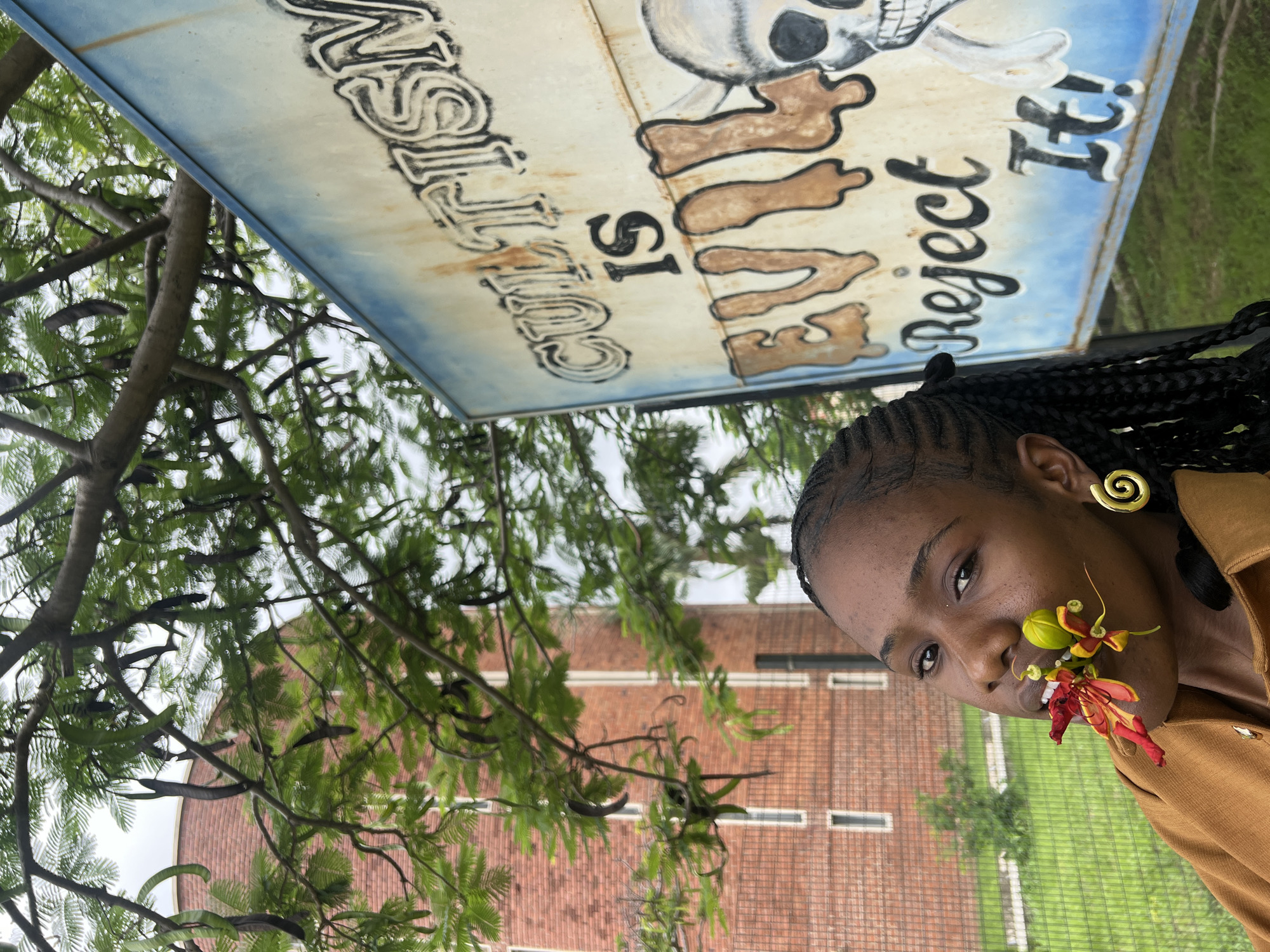 The image shows a young Black woman with braided hair and a golden spiral earring. She is looking directly at the camera with a neutral expression. She has a vibrant red and yellow flower tucked into the corner of her mouth. She is wearing a brown shirt.
Behind her, a sign with faded lettering is visible. The sign has a drawing of a skull and bone illustration. The text on the sign appears to read: "CUSTOM IS... EVIL! REJECT IT!". The sign is weathered and appears to be outdoors.
The background consists of lush green foliage from a tree and a red brick building with white window frames. The scene appears to be outdoors during the day, with overcast or cloudy weather suggested by the lighting. The overall impression is artistic and a bit rebellious, with the contrast between the woman's serene expression and the provocative message on the sign. The context of Jos, Nigeria, suggests a potentially urban or semi-urban setting.