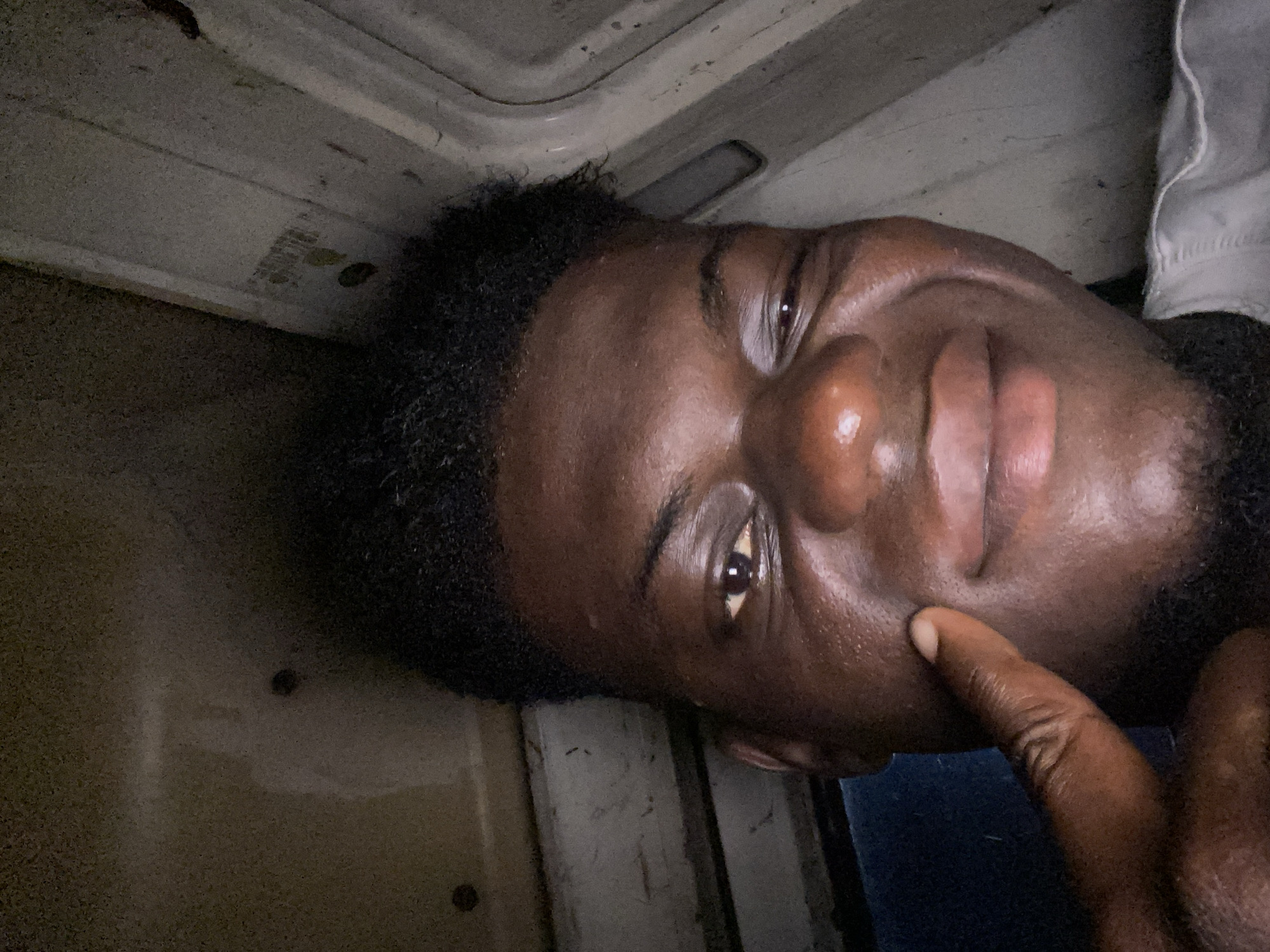 The image is a close-up portrait of a person, likely a young man, with dark skin and short, dark, curly hair. He is positioned at an unusual angle, with his head tilted, looking towards the viewer with a slight smile or smirk. His right eye is clearly visible and reflects light, while his right index finger touches his cheek in a seemingly thoughtful or playful gesture.
The background consists of light-colored, textured surfaces, possibly white or off-white, consistent with an enclosed, utilitarian space like the interior ceiling or overhead compartment of a vehicle (e.g., a van, bus, or train). Faint lines and patterns are visible on this surface, along with a small, rectangular light fixture in the upper portion of the frame. Part of a white garment is visible near the person's right shoulder, and a blue fabric is seen below his chin. The lighting is dim, suggesting an indoor or low-light setting. There is faint, illegible text present on the background surface in one of the zoomed-in crops. The person's expression conveys a sense of casualness or contentment.