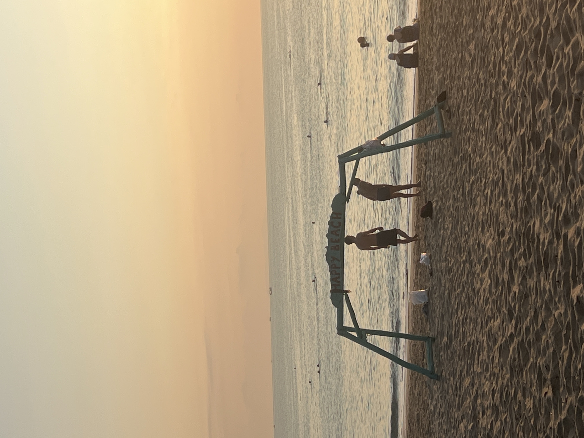 The image depicts a tranquil beach scene at what appears to be either sunrise or sunset, characterized by a warm, golden glow across the sky. The sun is not directly visible but casts a soft, diffused light, creating silhouettes of people and objects.
In the foreground, a sandy beach occupies the right side of the frame, showing textured patterns from footprints or waves. To the left, the calm, vast expanse of the sea stretches towards the horizon. Several people are visible, mostly in silhouette. Two figures are prominent near a green wooden structure positioned at the water's edge. One person stands directly behind the structure, facing the sea, while another stands slightly to the right, also looking towards the water. Further out in the shallow water, more indistinct figures can be seen, some appearing to be wading or standing. Several dark, small objects, possibly buoys or markers, dot the water surface in the mid-ground.
The green wooden structure acts as a sign, and the text "HAPPY BEACH" is clearly visible on its horizontal bar. This sign suggests a designated area or a local identifier for the beach. The overall atmosphere is peaceful and serene, typical of an early morning or late afternoon at the seaside. Given the context, this scene is set in Nha Trang, Vietnam.
