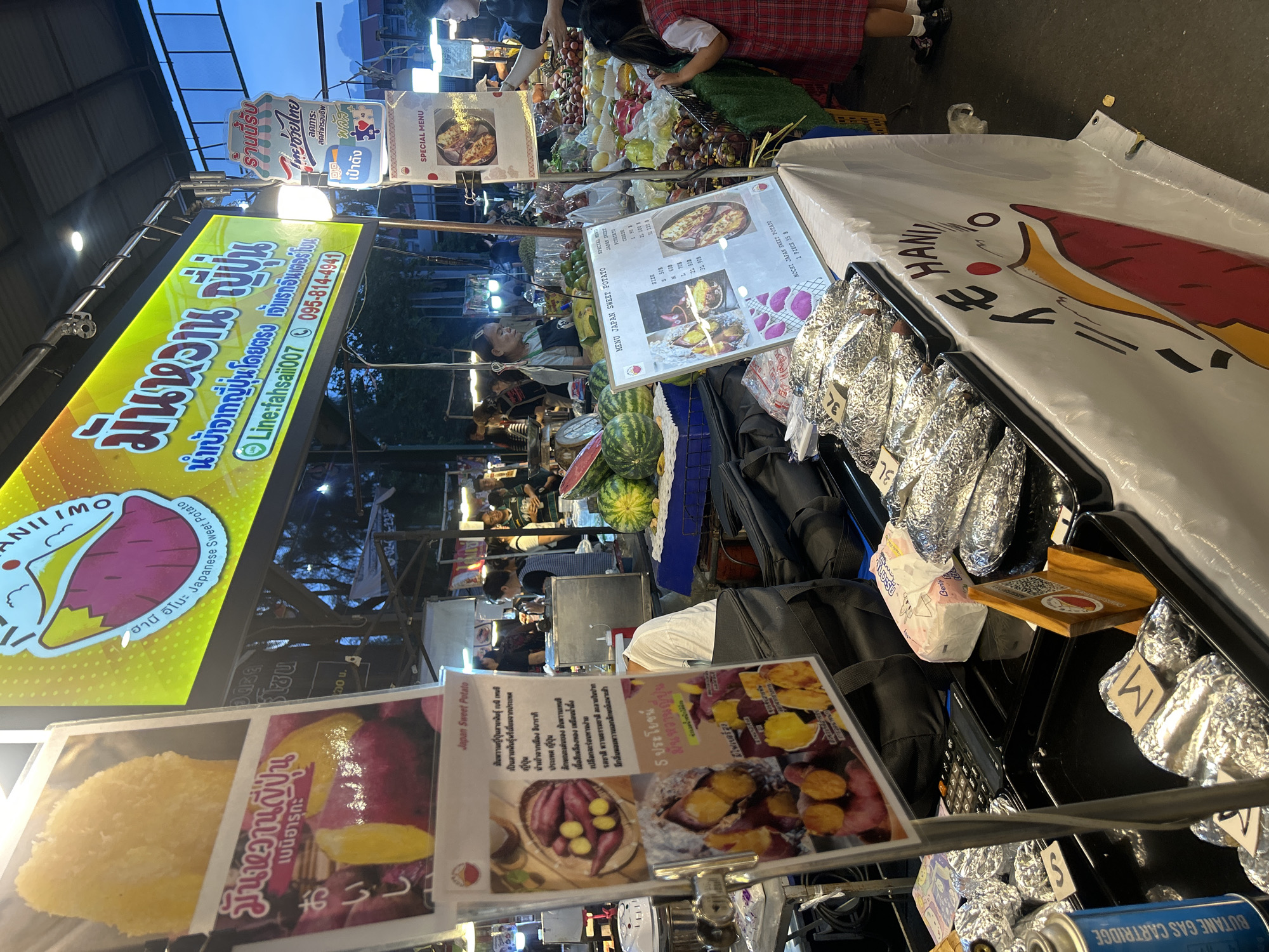 The image depicts an outdoor market stall in Tha Khlong Town Municipality, Thailand, specializing in Japanese sweet potatoes. A large, brightly lit yellow banner prominently displays text and an illustration of a sweet potato, indicating the name of the stall and contact information. Several signs in Thai and English advertise the products, including "Japan Sweet Potato" and "Special Menu."
The stall features various offerings, including whole sweet potatoes wrapped in foil, arranged in trays. Some signs display images of prepared sweet potato dishes, such as baked sweet potatoes and sweet potato slices. A young girl in a red and white checkered dress stands near the fruit stall, observing the merchandise. Other individuals are present, engaged in activities related to the market, such as attending to customers or preparing food.
The environment suggests a lively market atmosphere, with other stalls visible in the background selling fruits and other food items. The lighting indicates it is either late afternoon or early evening. The overall scene captures a snapshot of a typical Thai market, with a focus on a specific food vendor.