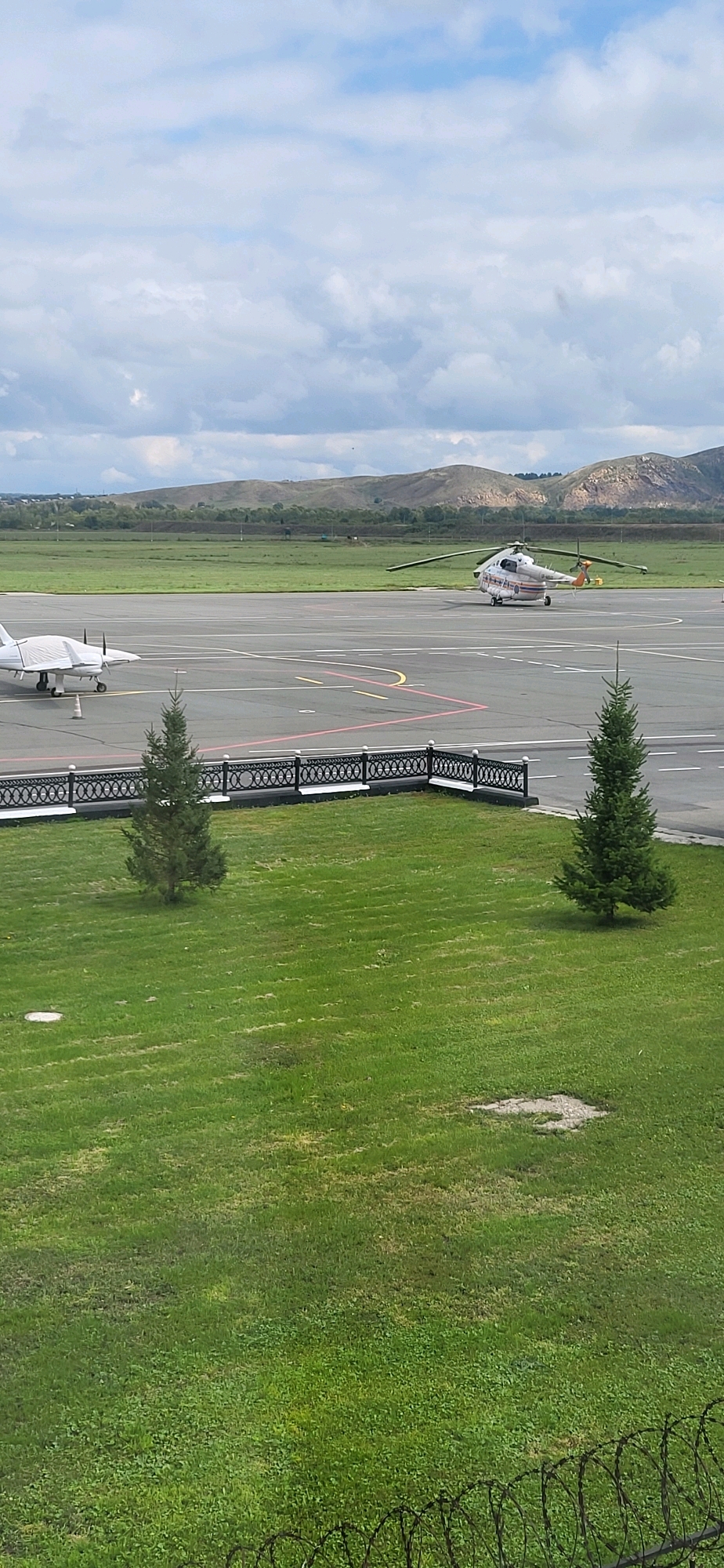 The image depicts an outdoor scene at an airport, likely located in Oskemen, Kazakhstan. In the foreground, a well-maintained grassy area is visible, bordered by a decorative black metal fence with white detailing. Two young evergreen trees stand on the grass. Beyond the fence, an asphalt tarmac is present, marked with yellow and white lines.
On the tarmac, two aircraft are visible. To the left, a small white airplane is partially covered with a protective tarp, with its propeller and wing clearly visible. A small traffic cone is placed near its front wheel. To the right, a larger, white helicopter with orange and grey markings is positioned on the tarmac, its blades extended. The helicopter appears to be operational.
In the background, a flat landscape unfolds, featuring green fields and rolling hills under a partly cloudy sky. The overall impression is one of a functional, possibly regional, airport environment. There are no people visible in the image.