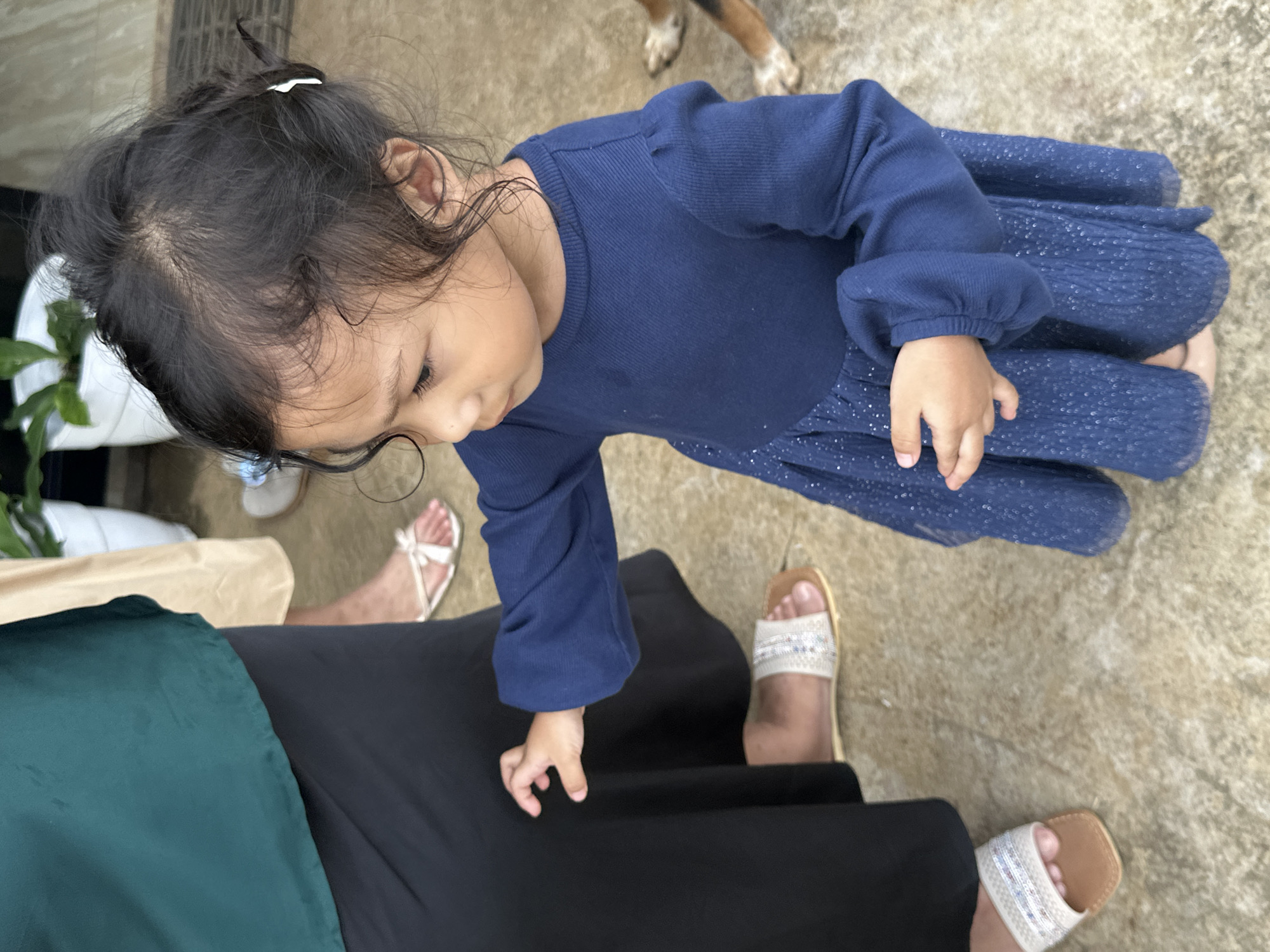 In Davao City, Philippines, a young child in a blue dress reaches out towards a black skirt worn by another person. Several pairs of sandals are visible on the ground, indicating an outdoor setting. The child's hair is dark and curly, with a white clip in the back. Part of a dog's paw is visible in the background. The ground appears to be paved with concrete tiles.