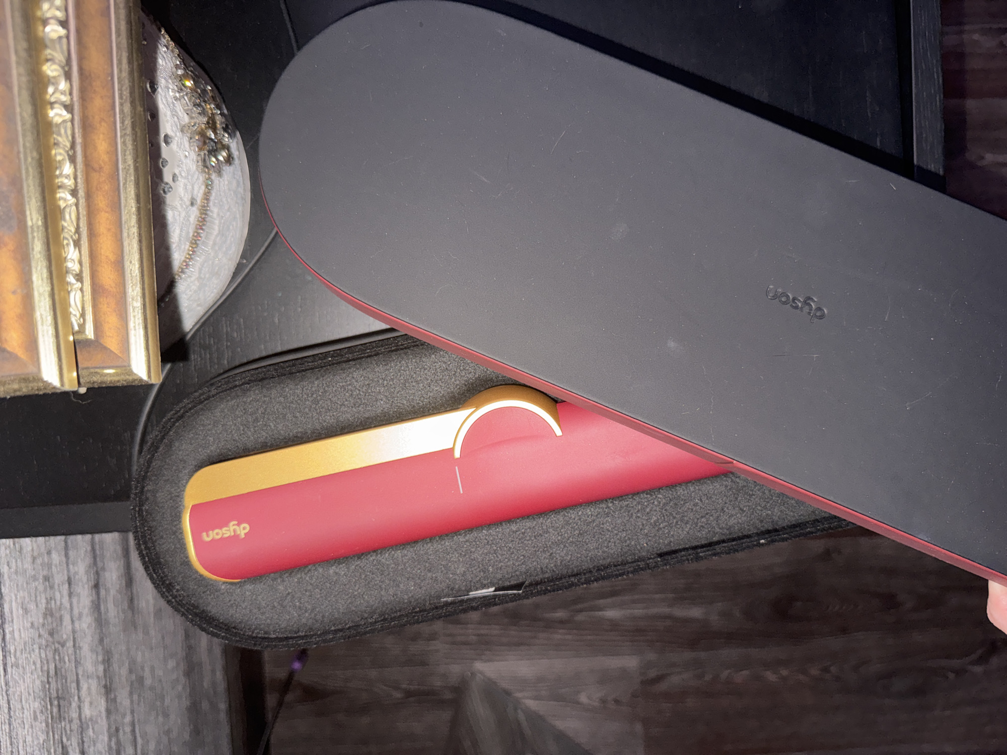 A red and gold Dyson straightener is partially visible inside a black protective case. The case is open, revealing the straightener's pink body and gold accents. The straightener appears to be made of plastic and metal. The background consists of a dark wooden surface and a black table. A portion of an ornate, gold-trimmed picture frame is visible at the top of the image. The scene is lit by an overhead light source, casting shadows. There are no people or animals present. The location is not specified.