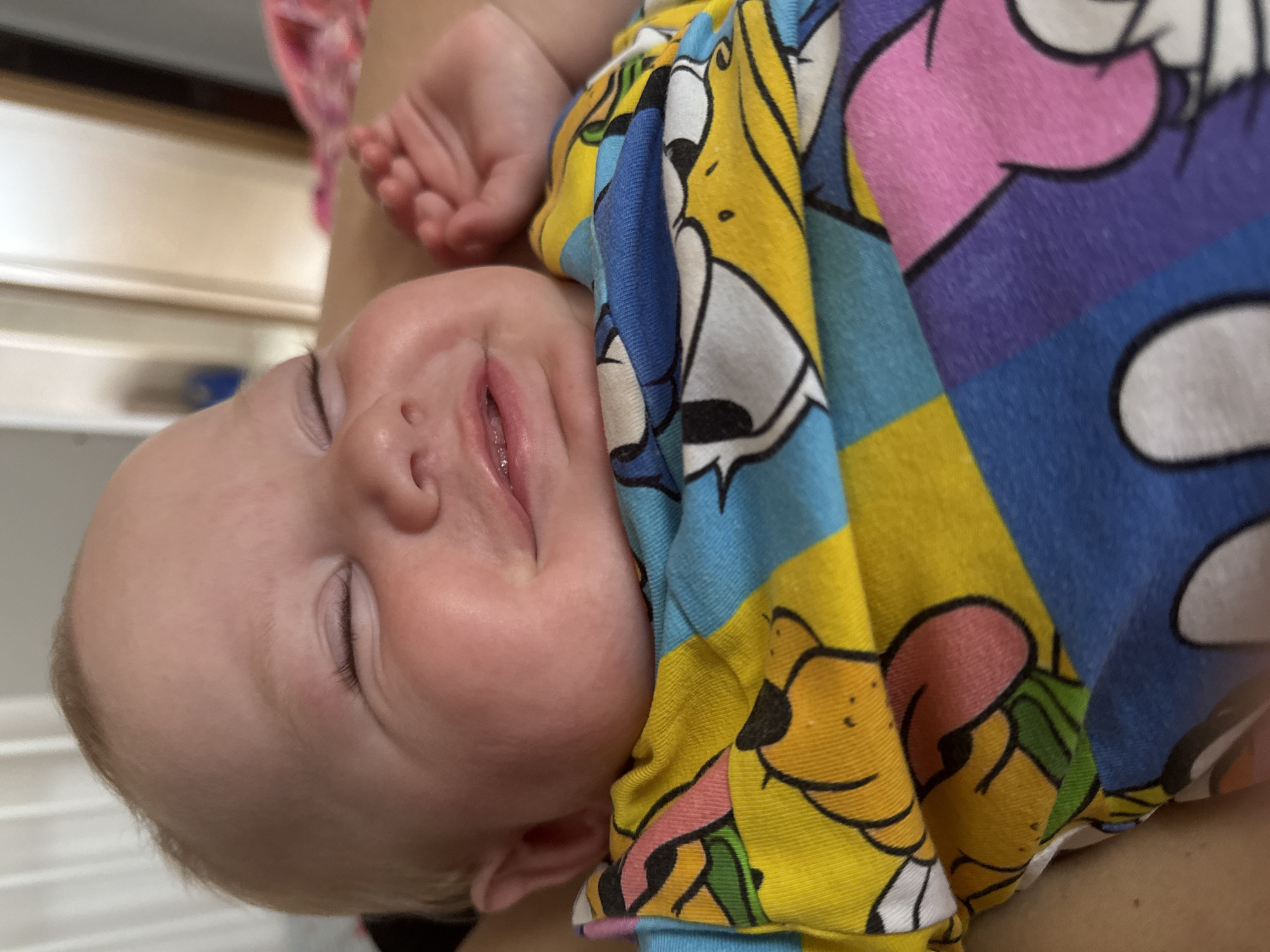 A light-skinned infant with fine, light-colored hair is depicted in a close-up, chest-up shot. The infant's eyes are closed, and its mouth is slightly open in a wide smile, revealing a wet lower lip and a hint of a small upper gum. The infant is wearing a vibrant, multi-colored garment, likely a t-shirt or onesie, adorned with various cartoon characters. Distinct sections show a yellow segment with the face of a character resembling Pluto, a blue segment featuring Donald Duck's head, and a purple-pink segment with a character resembling Minnie Mouse. Small, curled fists are partially visible on the infant's right side. The background is significantly blurred, suggesting an indoor setting with light-colored walls and possibly a door or furniture. The scene is situated in the city of Chaikovsky, Russia.