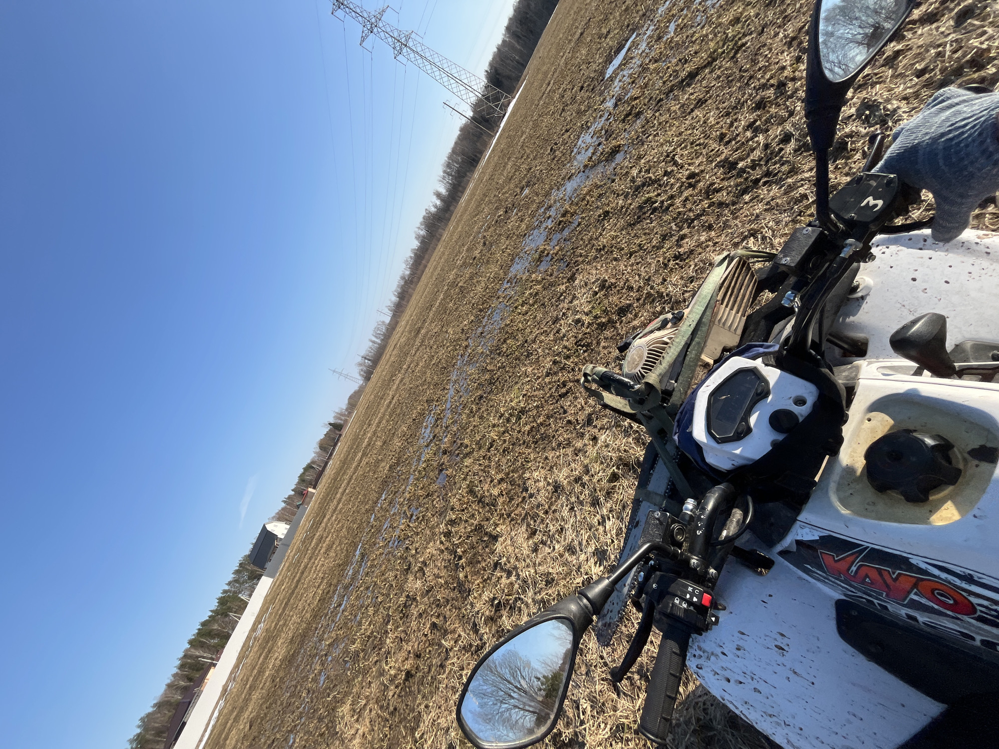 The image depicts a first-person perspective from a white all-terrain vehicle (ATV) in a muddy, open field in Razdol'e, Russia, likely during late winter or early spring under a clear blue sky.

The foreground is dominated by the front section of the ATV, splattered with mud. Visible components include the handlebars with black rubber grips and control clusters, a white digital instrument panel, a black ribbed fuel tank cap, and a portion of the seat. The brand "KAYO" in red is visible on the side fairing. A blue/grey gloved hand rests on the right handlebar. A chainsaw, secured with straps, is mounted on the front rack behind the handlebars. The left rearview mirror reflects bare trees against the blue sky.

The middle ground consists of a vast, uneven field covered in dried, brown grass or crop stubble, interspersed with numerous puddles of standing water and small patches of melting snow.

In the distant background, a line of predominantly bare deciduous trees and some coniferous trees forms a horizon. Along this treeline, several residential-style buildings are visible, including a prominent white house with a dark roof. A tall white fence runs parallel to a section of the treeline. To the far right, a large metal lattice electrical transmission tower stands prominently, with power lines extending horizontally across the frame. Another, smaller transmission pole is visible further in the distance. The sky above is uniformly clear and bright blue.