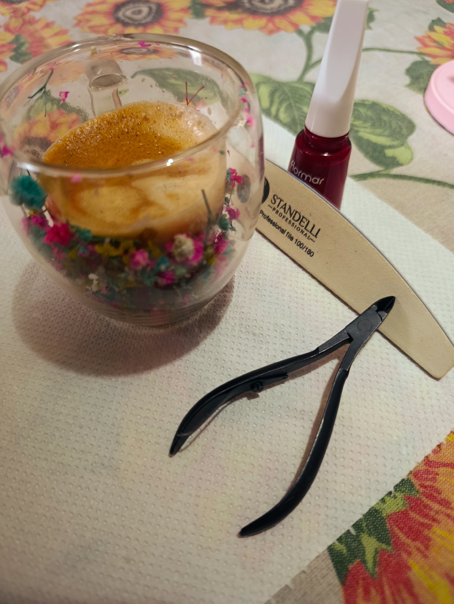 The image depicts a still life of nail care items on a surface covered with a floral patterned cloth. A clear glass containing what appears to be a drink with colorful decorative flowers surrounding it sits to the left. Adjacent to the glass, a red nail polish bottle stands next to a nail file labeled "STANDELLI PROFESSIONAL File 100/180". Below these items are black cuticle nippers.
There is no evidence to determine if the picture was taken in Kardzhali, Bulgaria.