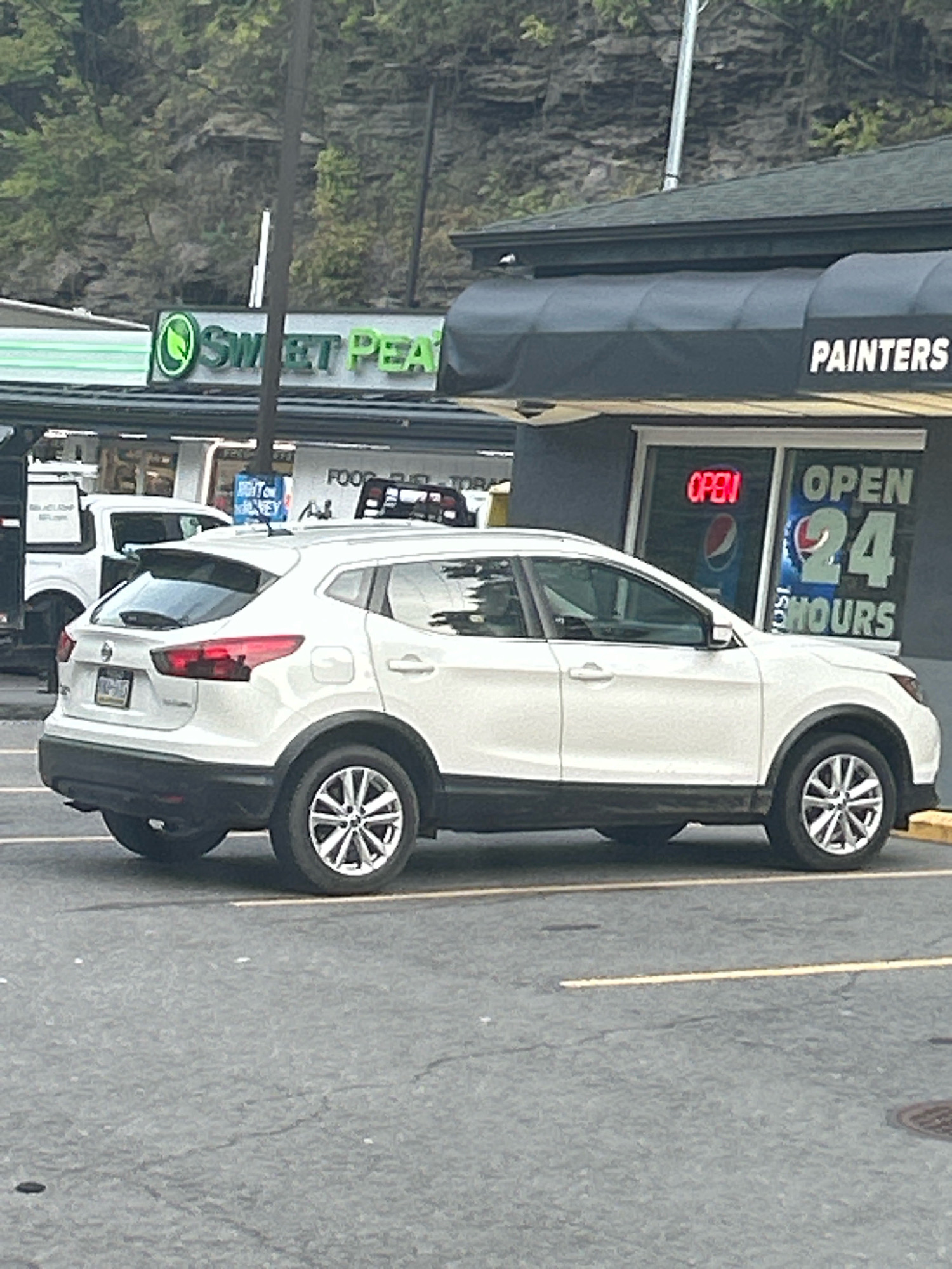A white Nissan Rogue is parked in a lot in front of a building. The building has a dark gray awning with "PAINTERS" in white lettering above a storefront. A sign in the window of the storefront reads "OPEN 24 HOURS" with a Pepsi logo visible. To the left of the building is a sign for "SWEET PEA." In the background, a wooded hillside is visible. The scene appears to be a commercial area. The location is not specified beyond being in the city of Pittsburgh, United States of America.