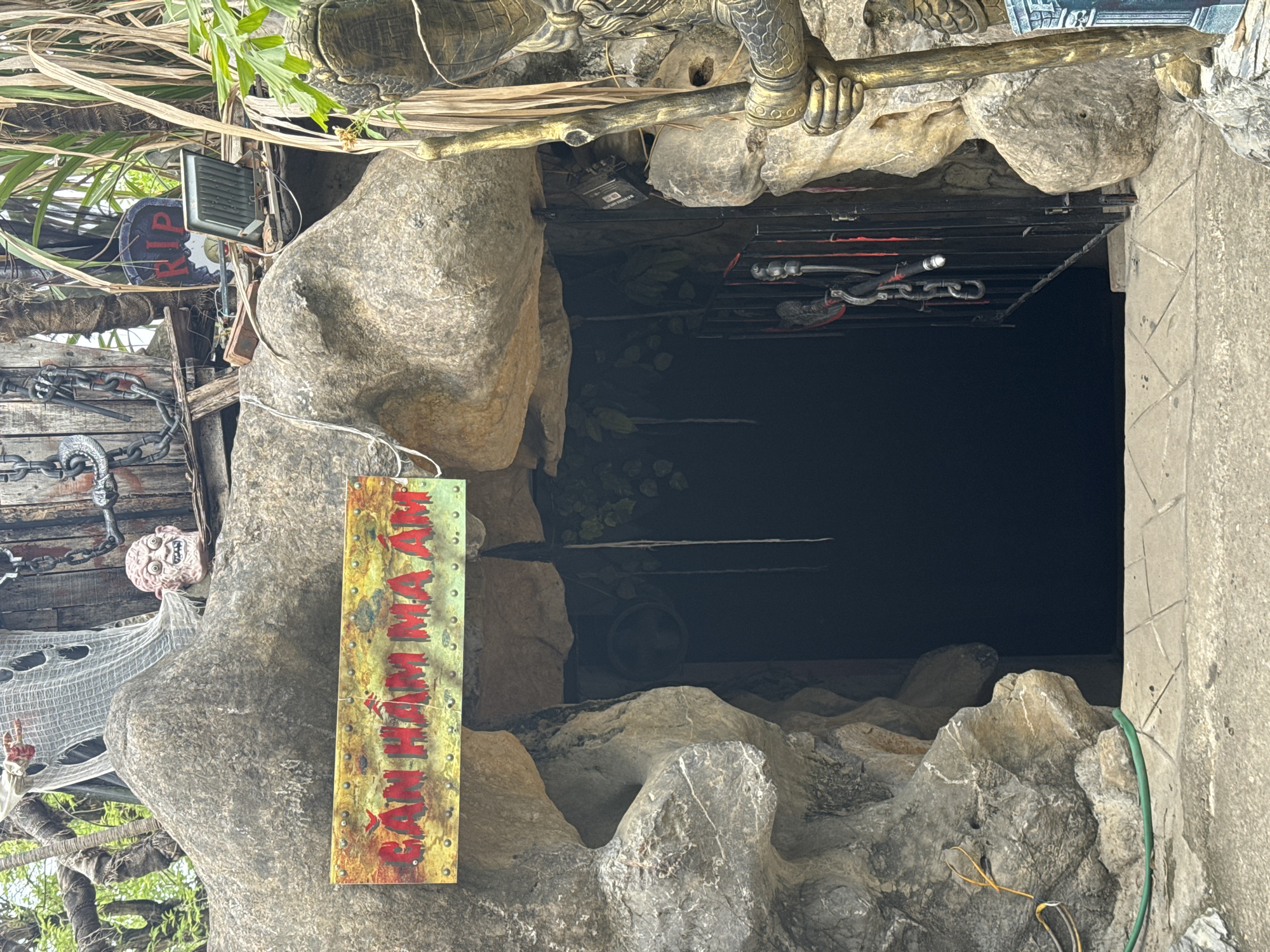 The image shows the entrance to a cave-like attraction, likely a haunted house or similar theme, in Hoa Lư Ward, Vietnam. A weathered sign above the entrance reads "CĂN HẦM MA ÁM" in red lettering against a distressed yellow background, translating to "Haunted Chamber." The entrance itself is a dark, cavernous opening framed by rough, grey rock formations.
Above the entrance, a macabre scene is depicted. A grotesque, disembodied head with wide eyes and an open mouth rests on the rocky overhang. Tattered white fabric, resembling cobwebs or spectral attire, drapes down from above, adding to the eerie atmosphere. A large, rusty chain hangs to the left, further enhancing the spooky theme. To the right, a tombstone-shaped sign with "R.I.P." in red letters is visible, along with a floodlight.
In the foreground, a statue of a warrior figure, elaborately decorated in gold and dark tones, stands to the right of the entrance. The statue holds a long staff. The ground leading to the entrance is paved with textured concrete tiles. Lush green foliage and palm fronds are visible in the background, suggesting an outdoor setting, possibly in a park or tourist area within Hoa Lư Ward. The overall impression is of a deliberately unsettling and theatrical environment designed to evoke fear and amusement.