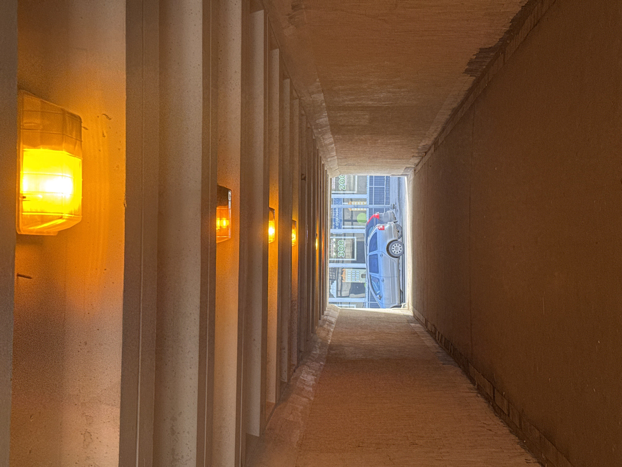 The image shows the interior of a pedestrian tunnel in Moscow, Russia. The tunnel has concrete walls with vertical ribs on the left and a smooth, tan wall on the right. Yellow lights are mounted on the ribbed wall. At the end of the tunnel, a cityscape is visible, including a gray minivan parked near a storefront with a sign. The overall lighting in the tunnel is dim, contrasting with the brighter daylight outside.