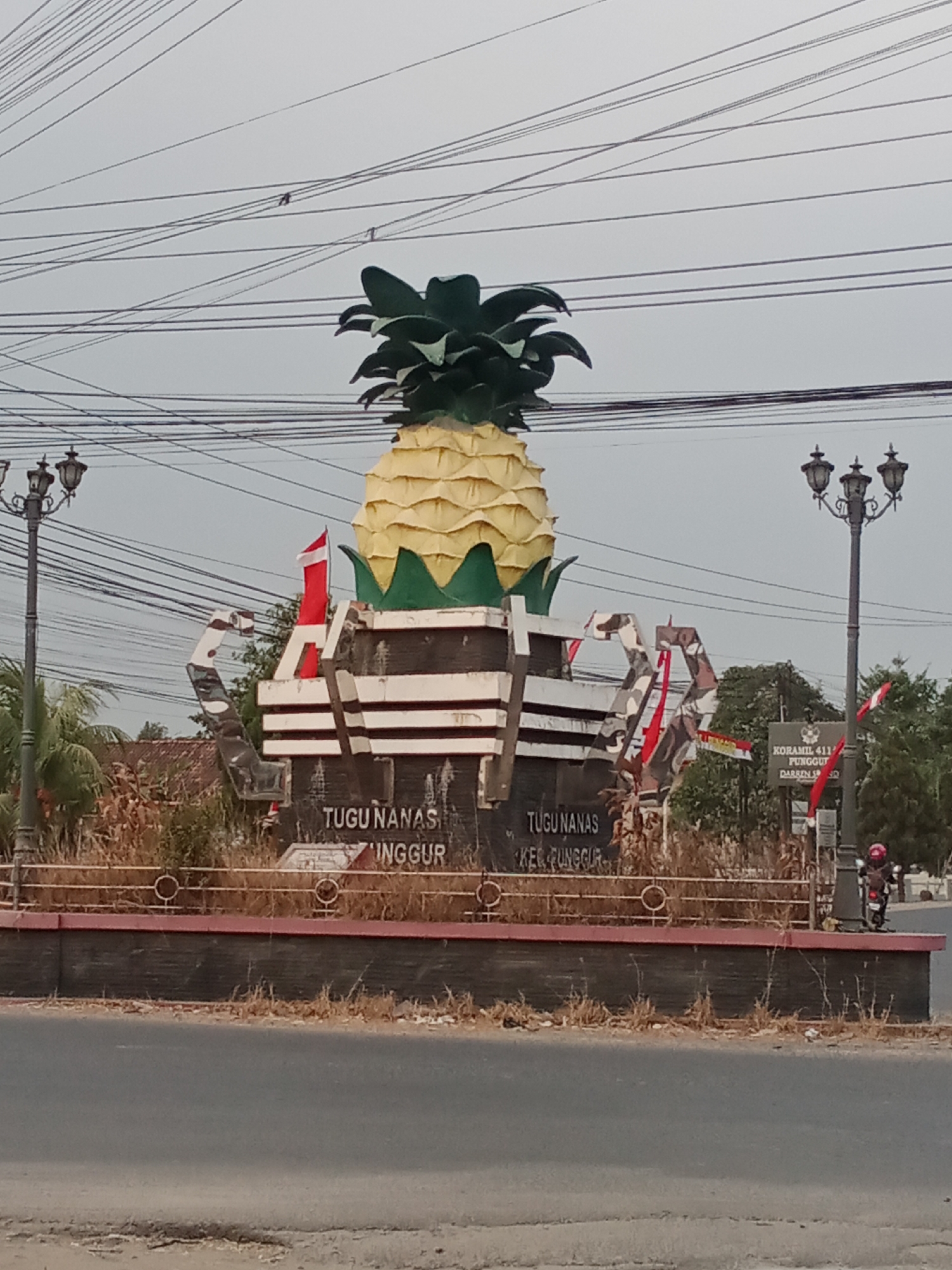 A monument in the shape of a large pineapple stands prominently in the foreground of the image, situated at the Tugu Nanas intersection in Toto Katon, Indonesia. The monument is constructed of concrete and painted in yellow and green, representing the fruit. The base of the monument features text that reads "TUGU NANAS KEC. PUNGGUR".
Flanking the monument are two ornate streetlights on either side, casting a dim light. Numerous power lines crisscross the sky above, creating a dense network. In the background, several buildings and trees are visible, suggesting a semi-urban or rural setting. A red and white flag, likely the Indonesian flag, is attached to the monument.
A person on a red motorcycle is partially visible on the right side of the image, near the streetlight. The overall scene depicts a roadside monument in a locale characterized by electrical infrastructure and a modest amount of vegetation. The sky is overcast, indicating a possibly cloudy day. The road in the foreground is asphalt, with some dry grasses and debris along its edge.
