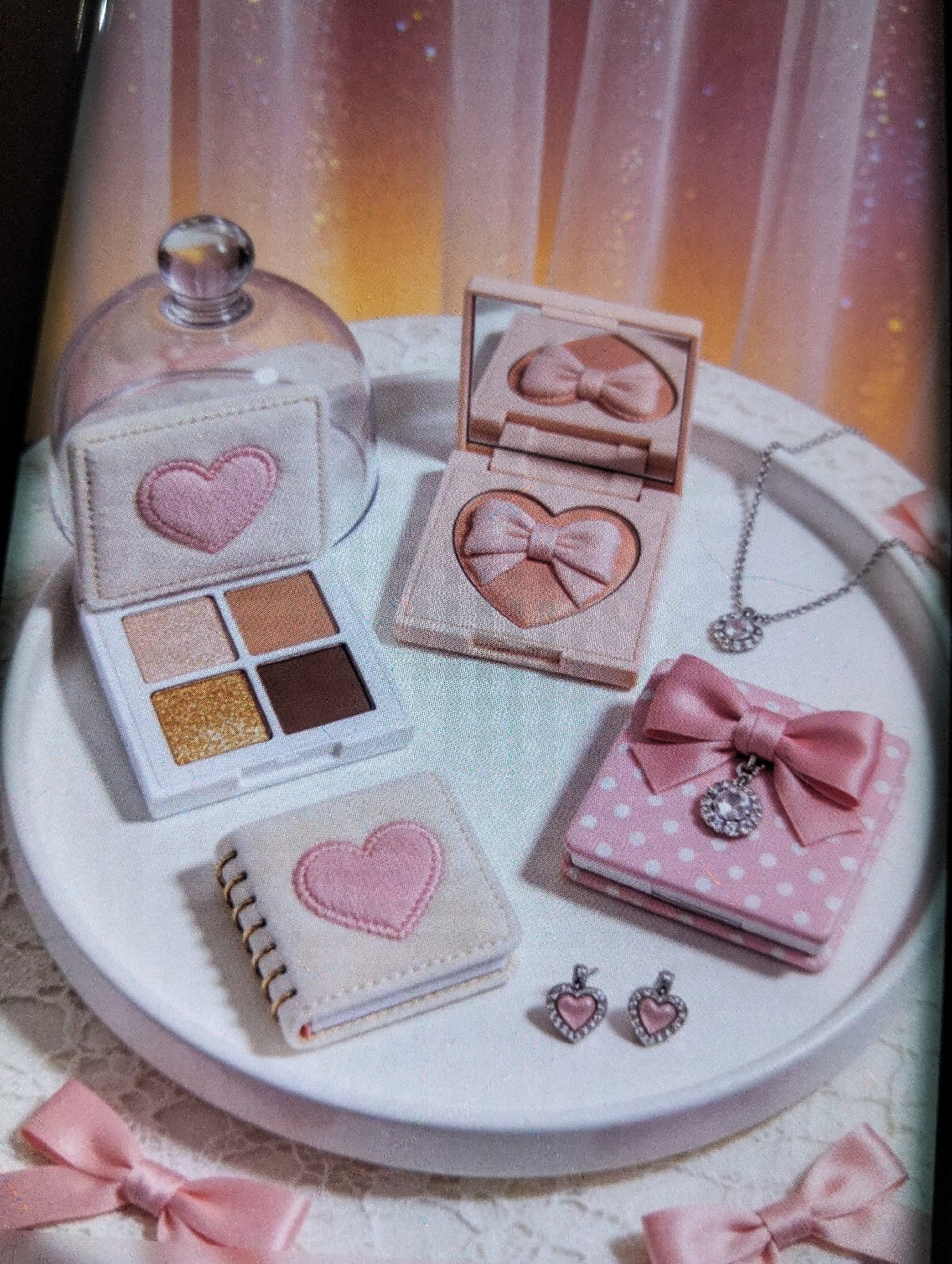 The image displays a collection of charming, heart-themed makeup and accessories arranged on a white circular tray. The items include an open eyeshadow palette with four shades, a compact blush or highlighter with a bow imprint, a small notebook with a heart design, a pink polka-dot box adorned with a bow and a pendant, a necklace with a pendant, and a pair of heart-shaped earrings. The overall aesthetic is soft and feminine, with a color palette dominated by pinks, peaches, and neutral tones, accentuated by sparkly elements and a subtle gradient background suggesting a diffused light source. There are no people or discernible elements of an urban environment like the city of Payagaboh, Indonesia in the image.