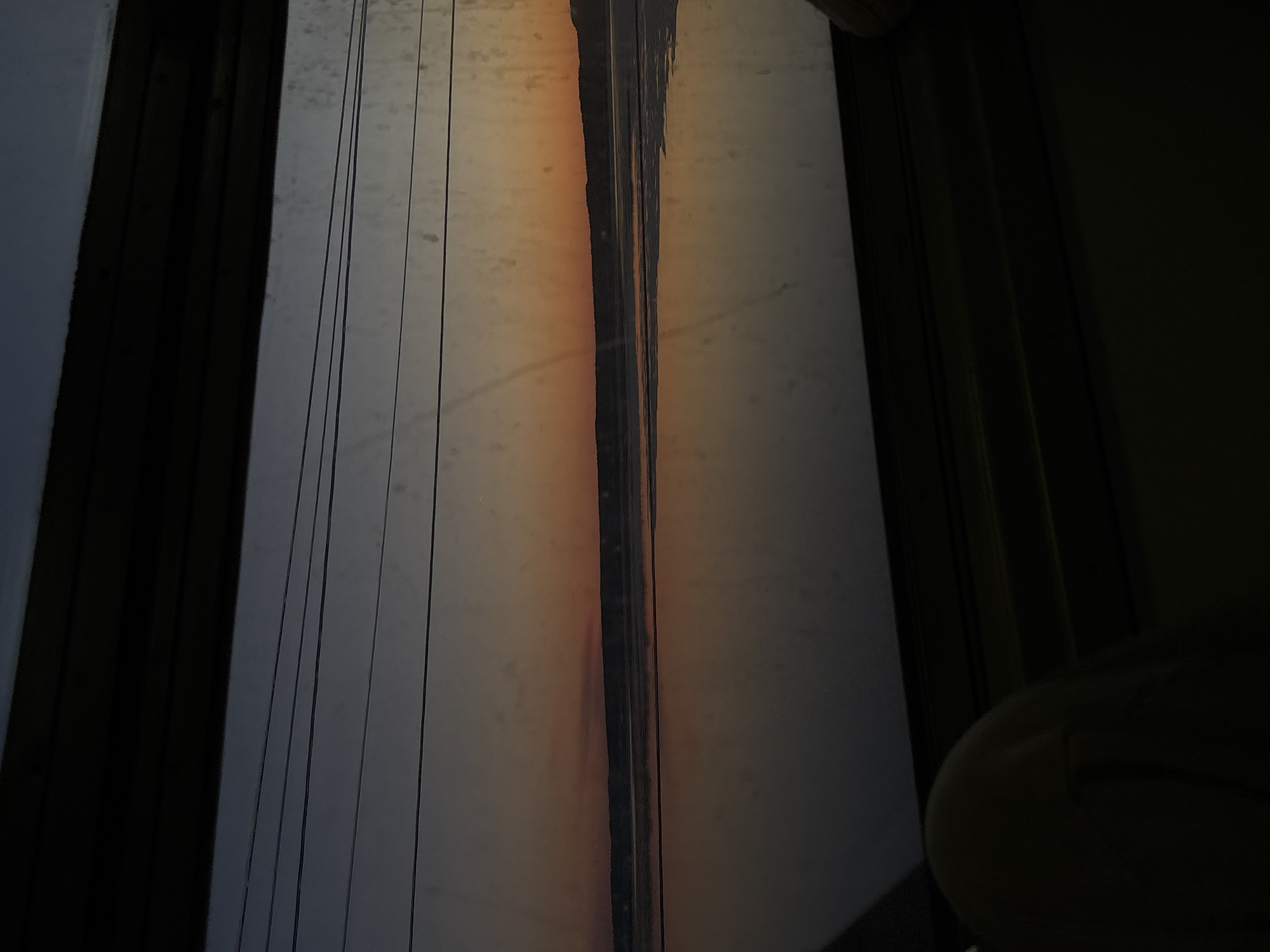 A view from inside a structure, potentially a building in Пальники, Russia, looking out through a window. The window is characterized by vertical lines, possibly wires or streaks on the glass. The light is somewhat diffused, creating an orange hue in the center of the window panes. The sides of the window frame are dark, with an obscured object or person in the lower right corner. The overall scene is dimly lit.