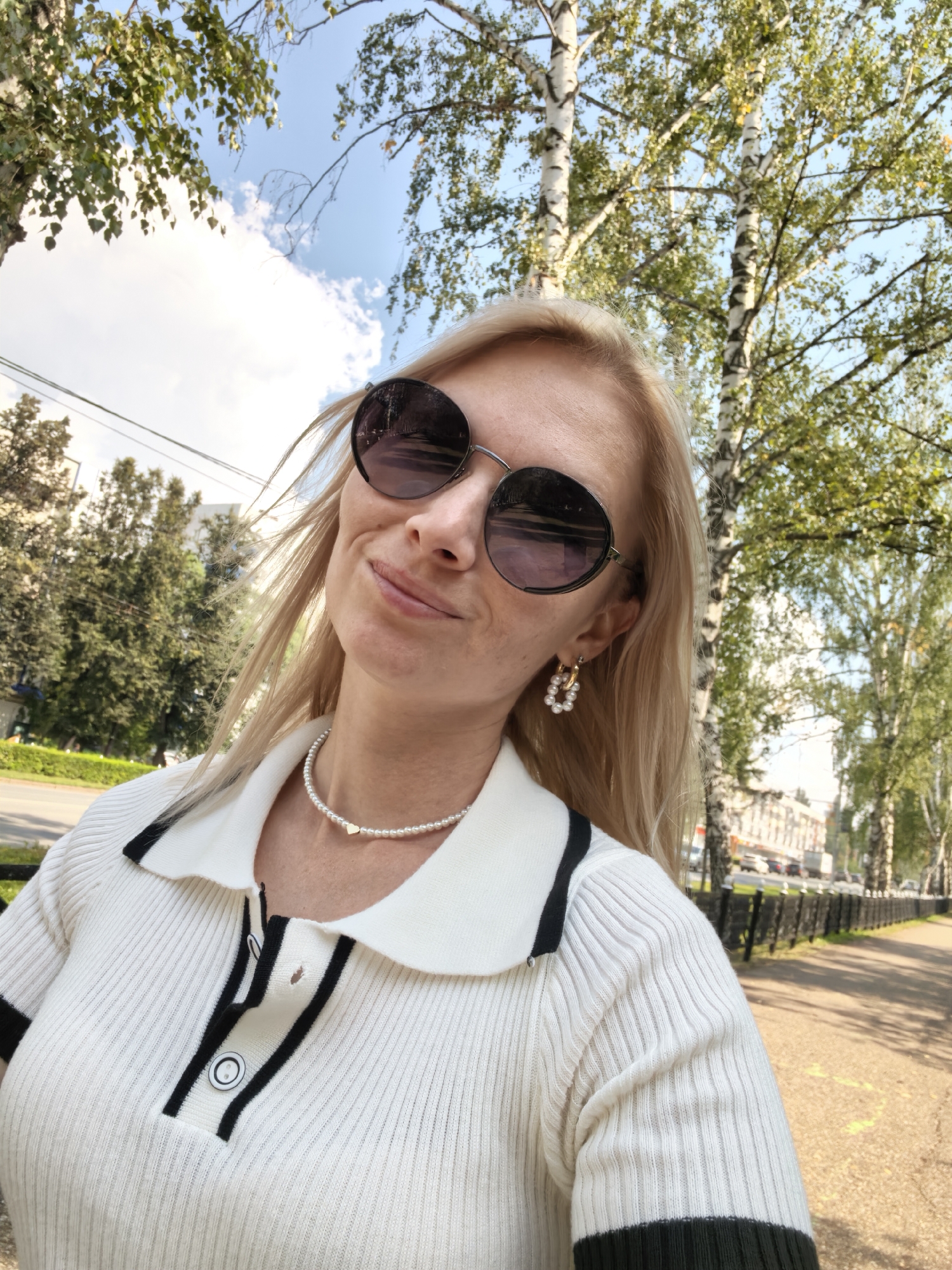 A blonde woman with sunglasses and a pearl necklace is taking a selfie outdoors on a sunny day. She is wearing a white ribbed polo shirt with black trim and buttons. Behind her, there are birch trees, a blue sky with white clouds, and a street with cars and buildings in the distance. The image appears to have been taken in the city of Sterlitamak, Russia, based on the context provided.